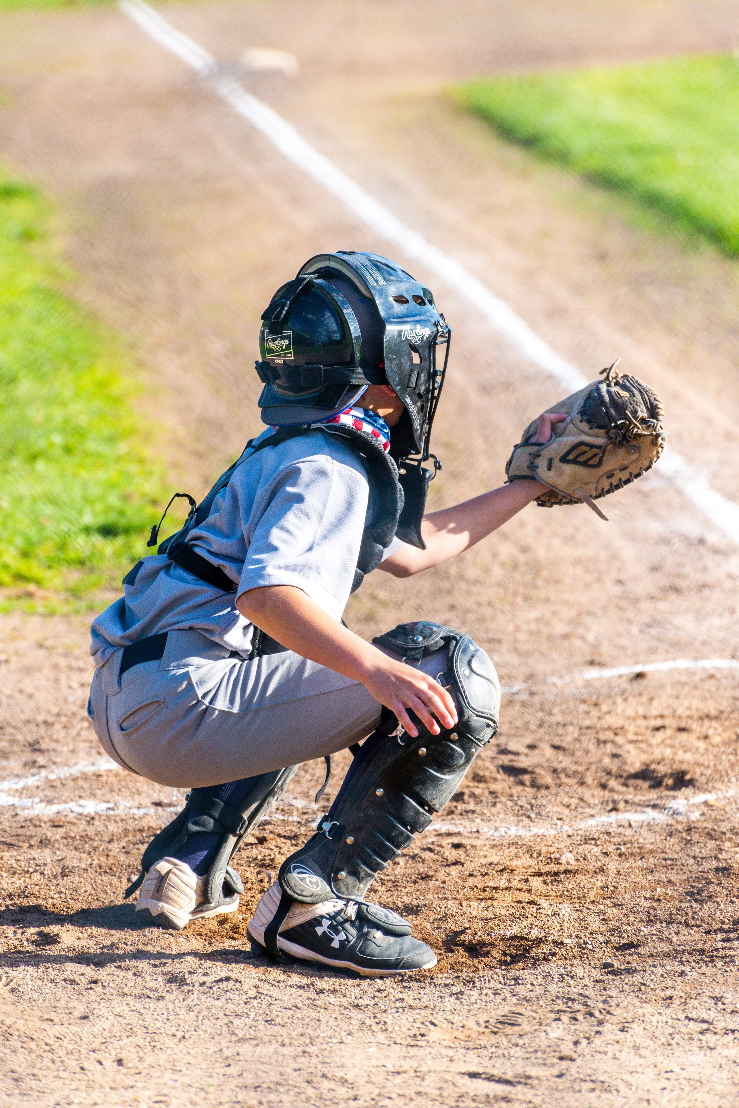A young baseball player in a catching stance, wearing a helmet and catcher's gear, on a dirt baseball field.
