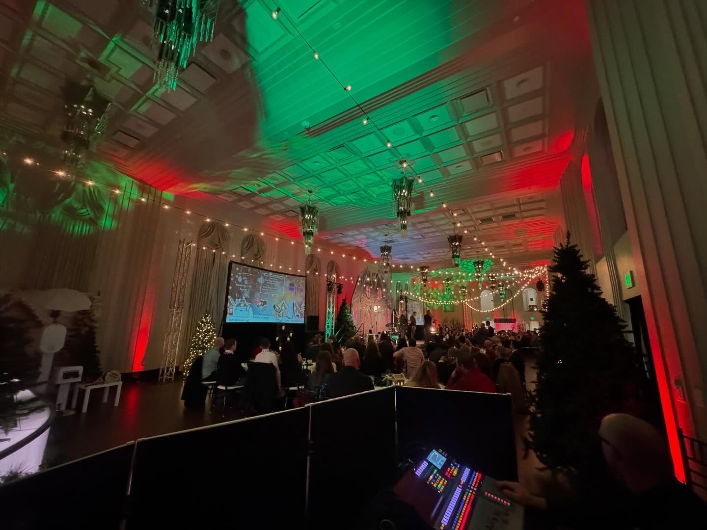 Indoor holiday party with Christmas trees, string lights, and a large crowd watching a stage performance in a decorated banquet hall.