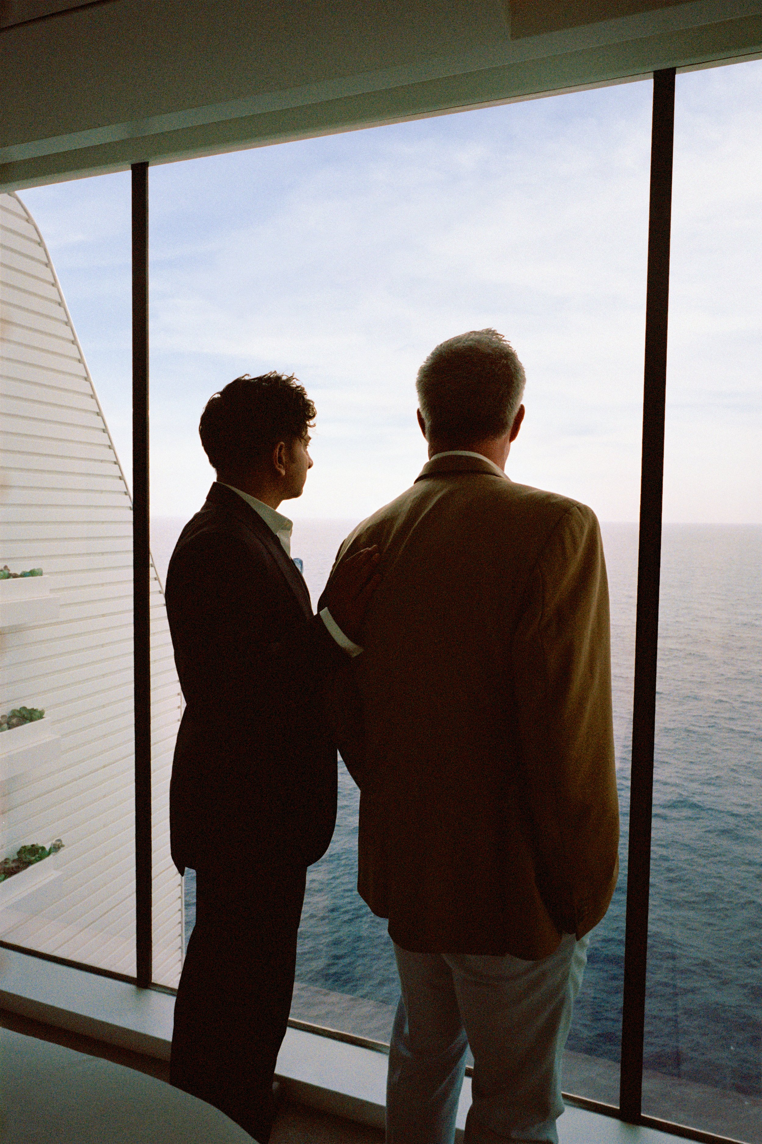 Two men in suits looking out a large window at the ocean on a cruise ship.