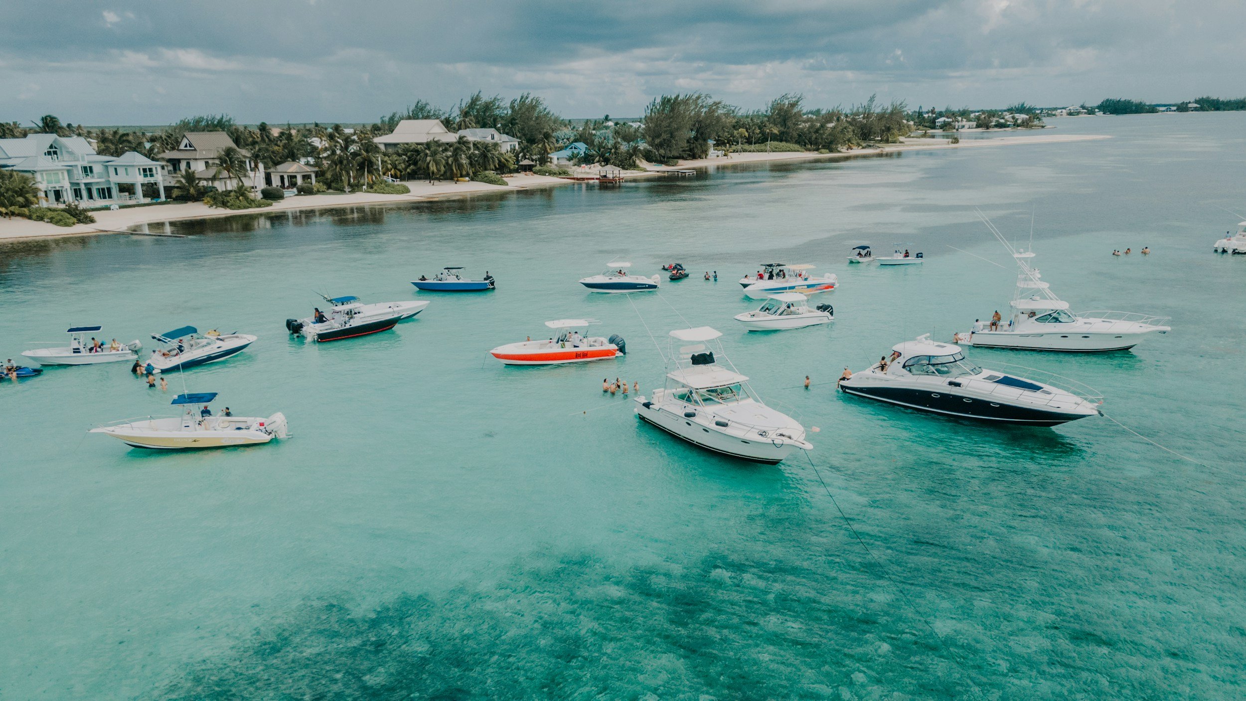 Multiple boats anchored in turquoise water near a shoreline with houses and palm trees.