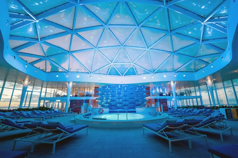 Indoor view of a modern, circular pool area inside a glass-domed structure with lounge chairs surrounding the pool.