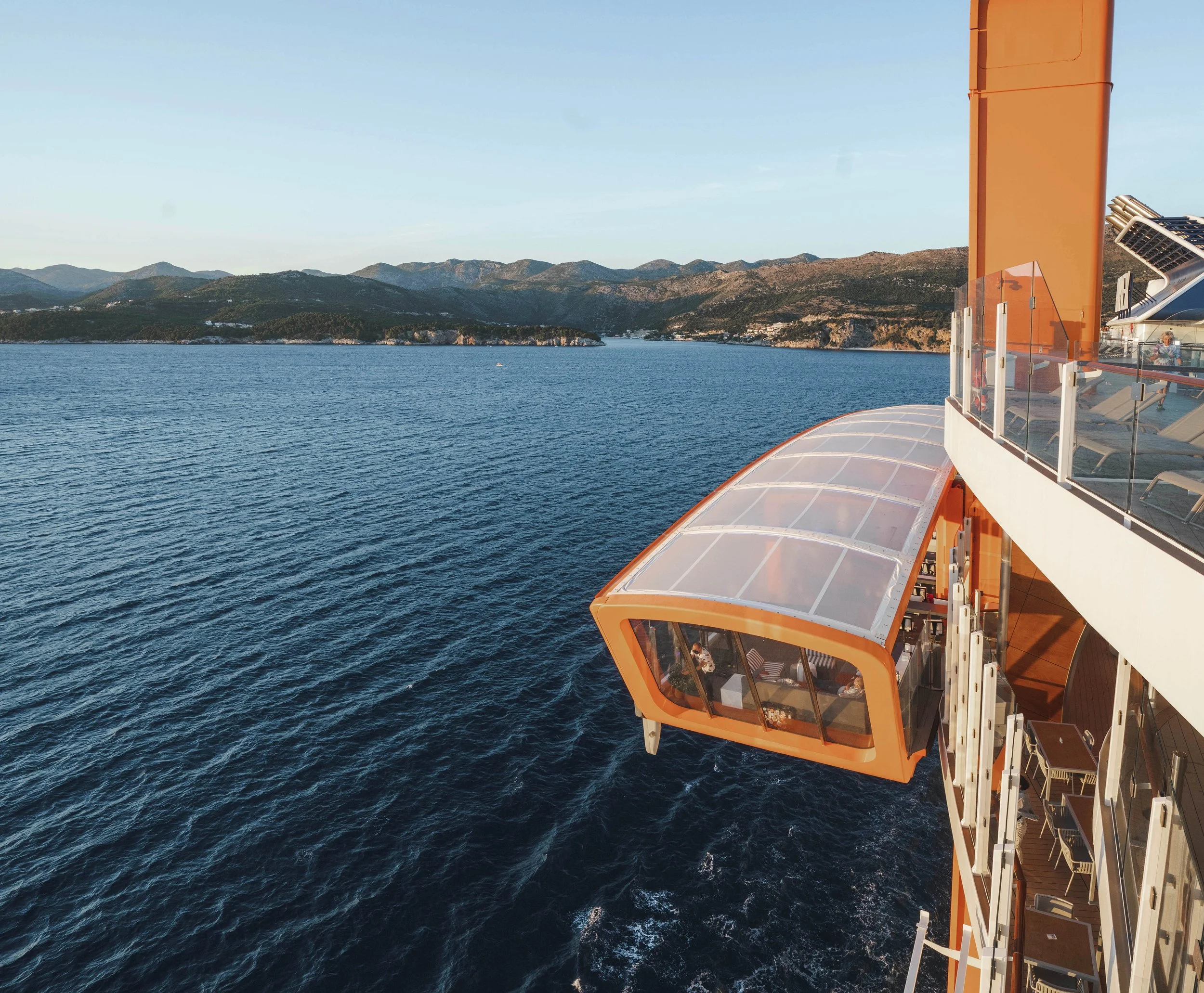 A february sunset on the water from a cruise ship observation deck with mountains in the background and glass-enclosed balcony with orange accents.