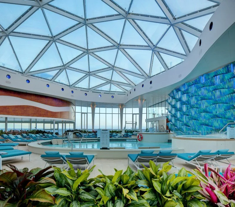 Indoor pool area on a cruise ship with lounge chairs, plants, and a glass-domed roof.