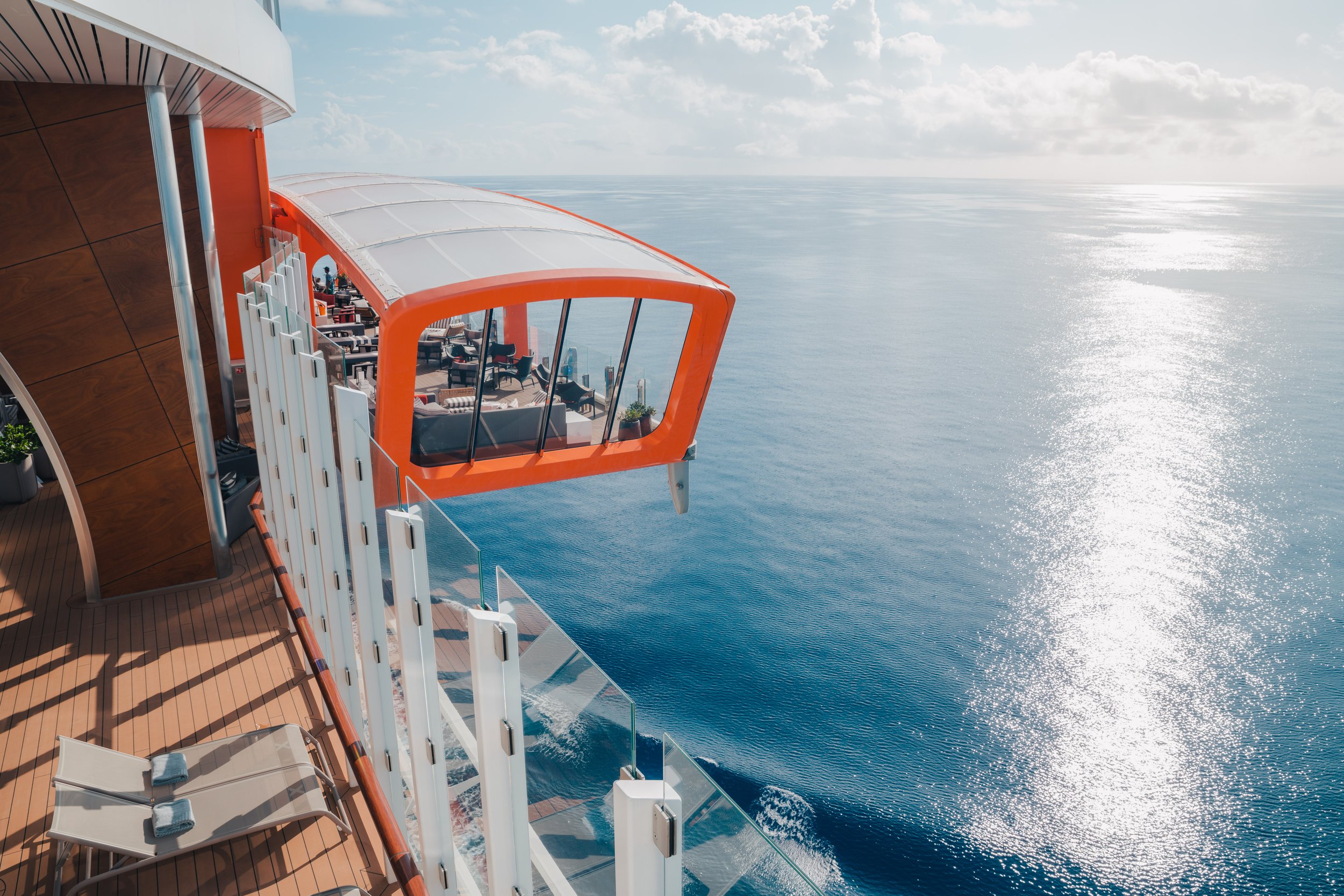 View from the deck of a cruise ship showing a glass-enclosed observation area with orange framing extending over the ocean, with blue sky and clouds in the background.