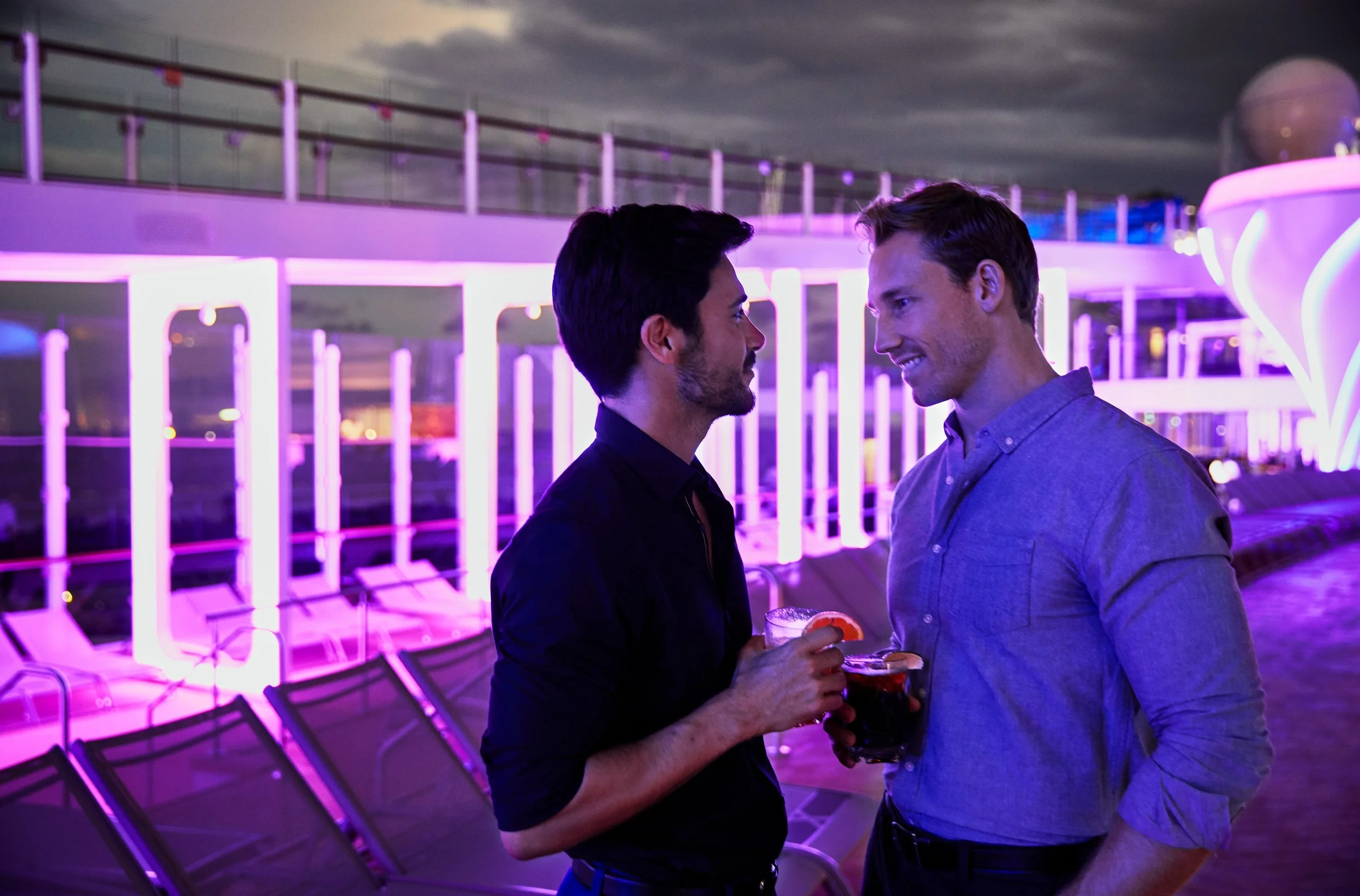 Two men are smiling and making eye contact at an outdoor event during evening or night time with purple and pink neon lighting. They are standing close to each other, holding drinks, with an empty row of chairs and a deck with glass railings in the background.