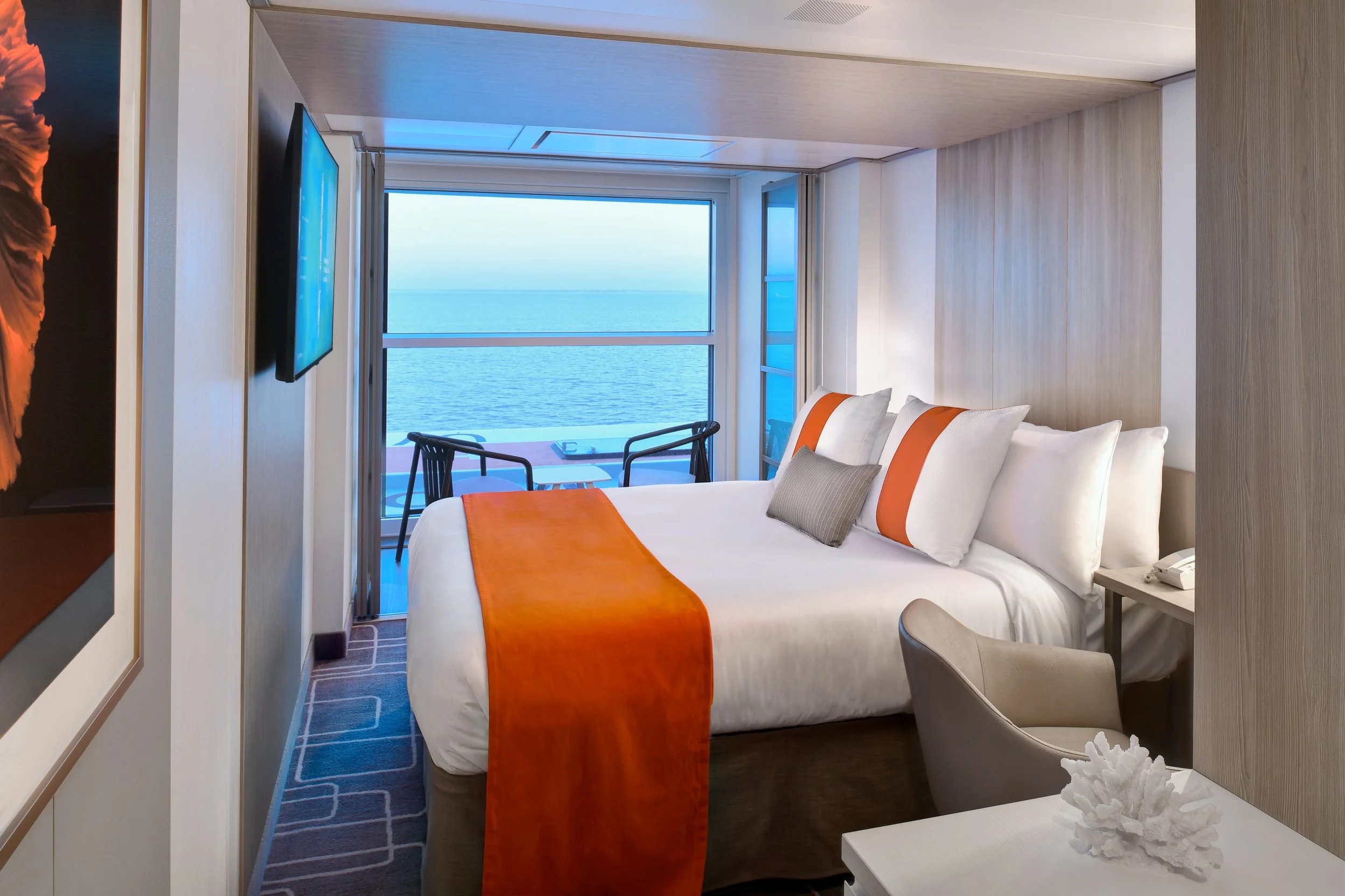 A hotel room with a view of the ocean through large glass sliding doors. The room has a bed with white linens and orange accents, a mounted flat-screen TV, a small sitting area, and a patterned carpet.