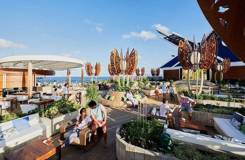 People relaxing and socializing on a rooftop lounge with stylish furniture, greenery, and decorative structures, with the ocean visible in the background under a clear blue sky.