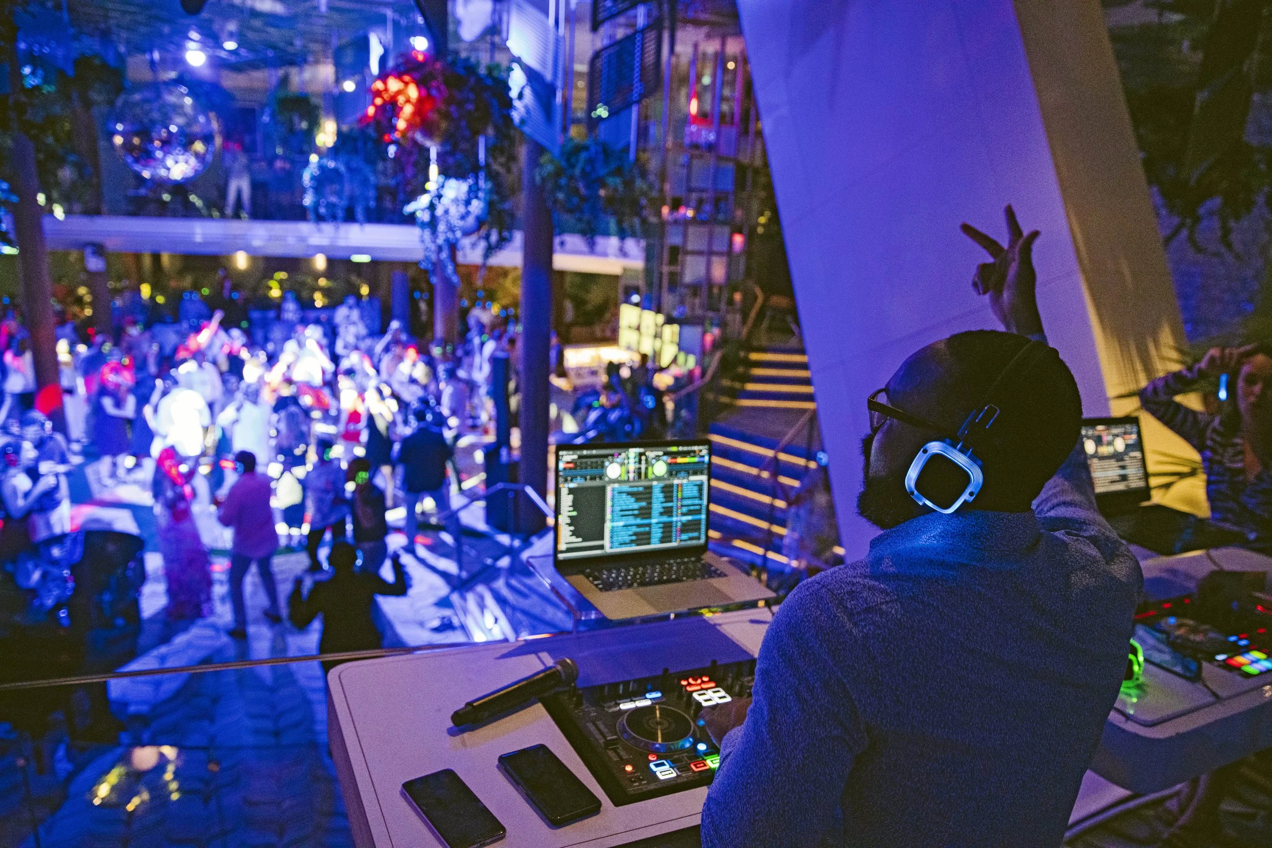 A DJ wearing glasses and headphones is working with a DJ controller and laptop, in a nightclub or event space, with a crowd dancing and socializing in the background illuminated by colorful lighting.