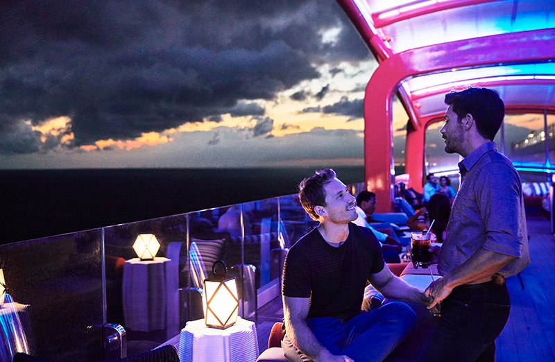 Two men having a conversation on a rooftop terrace during sunset, with a scenic view of the sky and clouds, colorful lighting, and seating with lanterns.