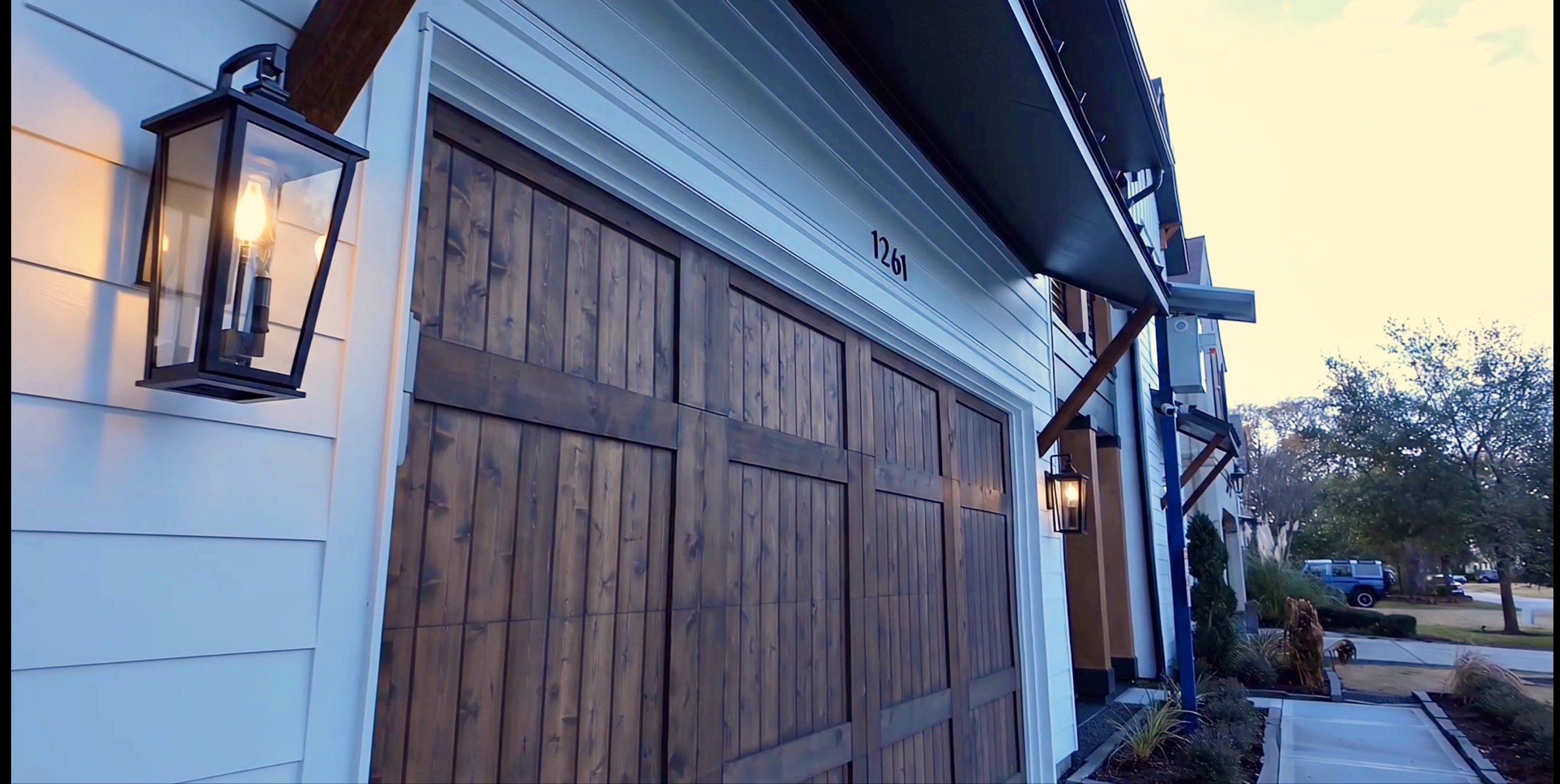 Exterior view of a modern apartment building with a wooden garage door, exterior lantern-style lights, and trees in the background during late afternoon or early evening.