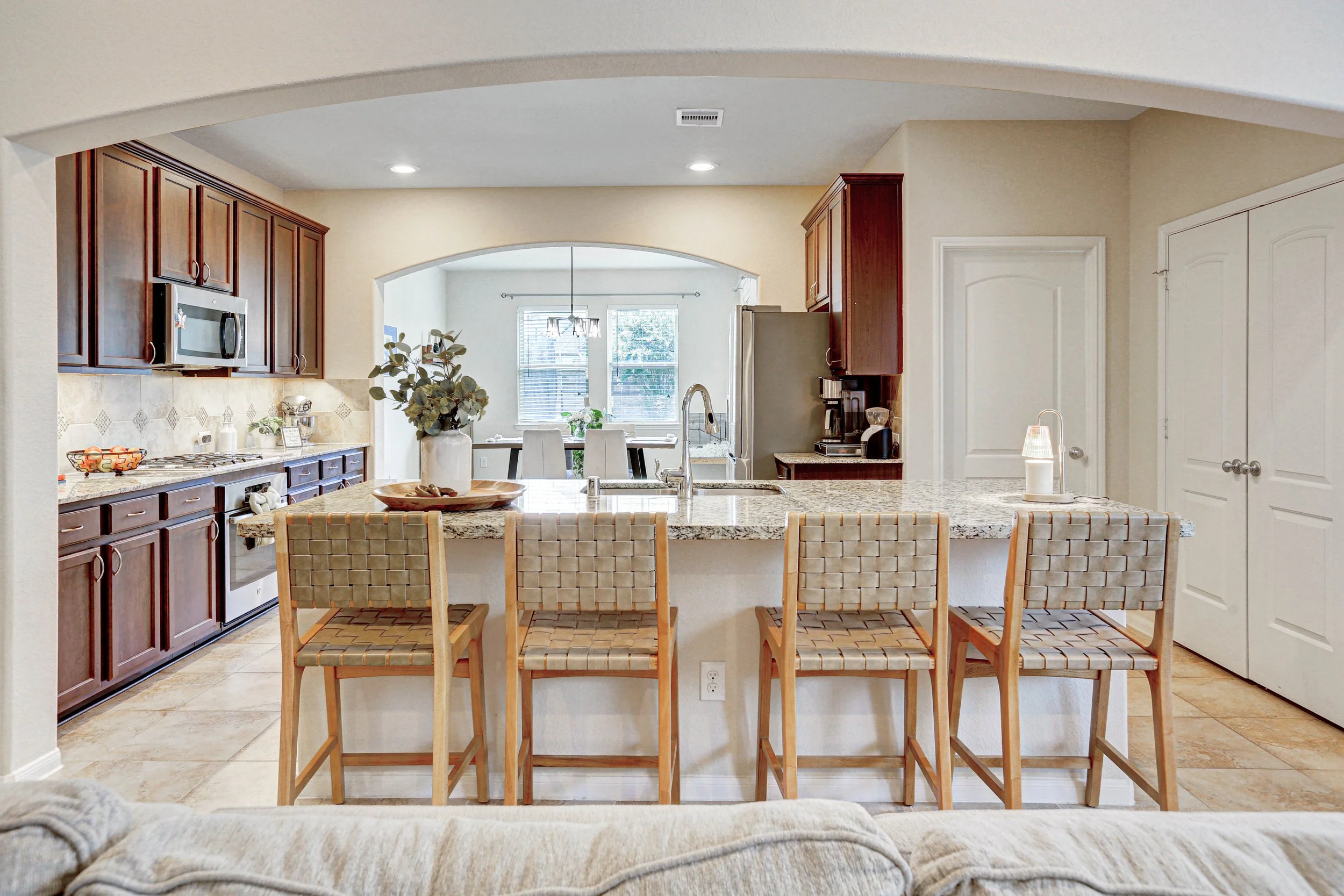 Open-concept kitchen with dark wood cabinets, granite countertops, and a breakfast bar with woven wood chairs. A side table with a lamp is visible in the foreground.