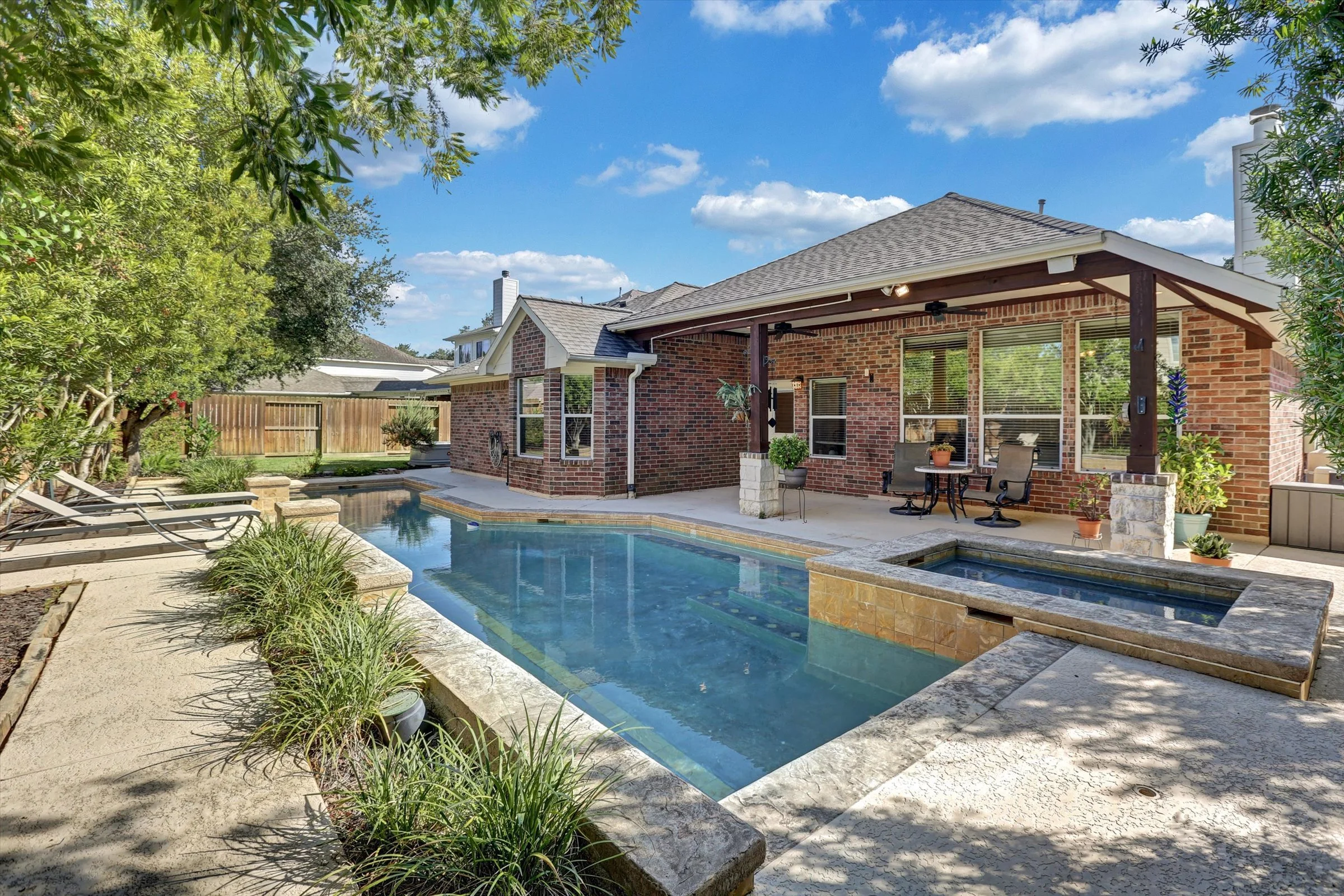 Backyard with swimming pool, patio furniture, and a fenced garden area with trees and plants, brick house with covered patio, and blue sky with clouds in Houston