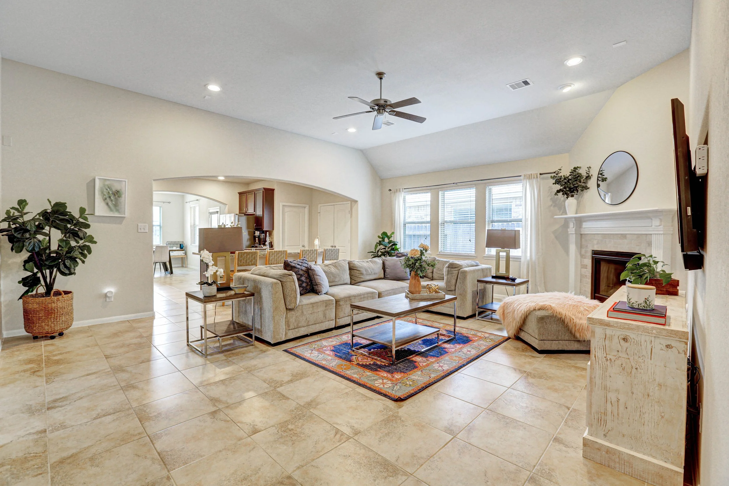 Spacious living room with beige tile floor, cream-colored walls, large windows with white curtains, a ceiling fan, and a cozy beige sectional sofa with decorative pillows. There are potted plants, a colorful area rug, and a fireplace with a round mirror above it.