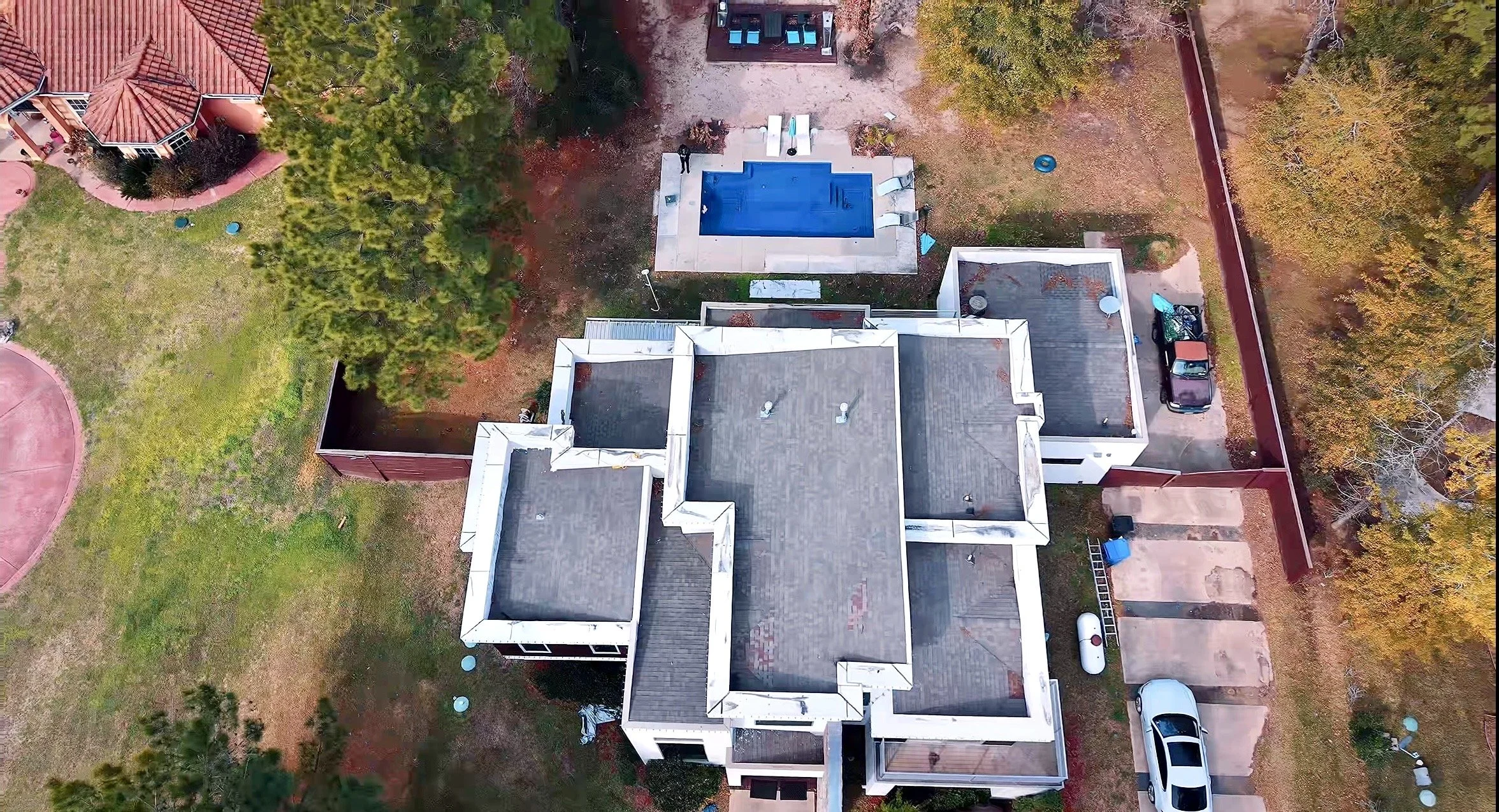 An aerial view of a residential backyard with a swimming pool, patio area, trees, a lawn, and parked cars along a fence in Houston