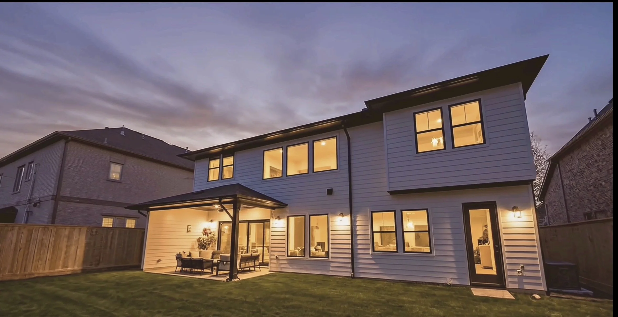 A modern two-story house with illuminated windows, a backyard with outdoor seating, and neighboring houses, during dusk with a cloudy sky.