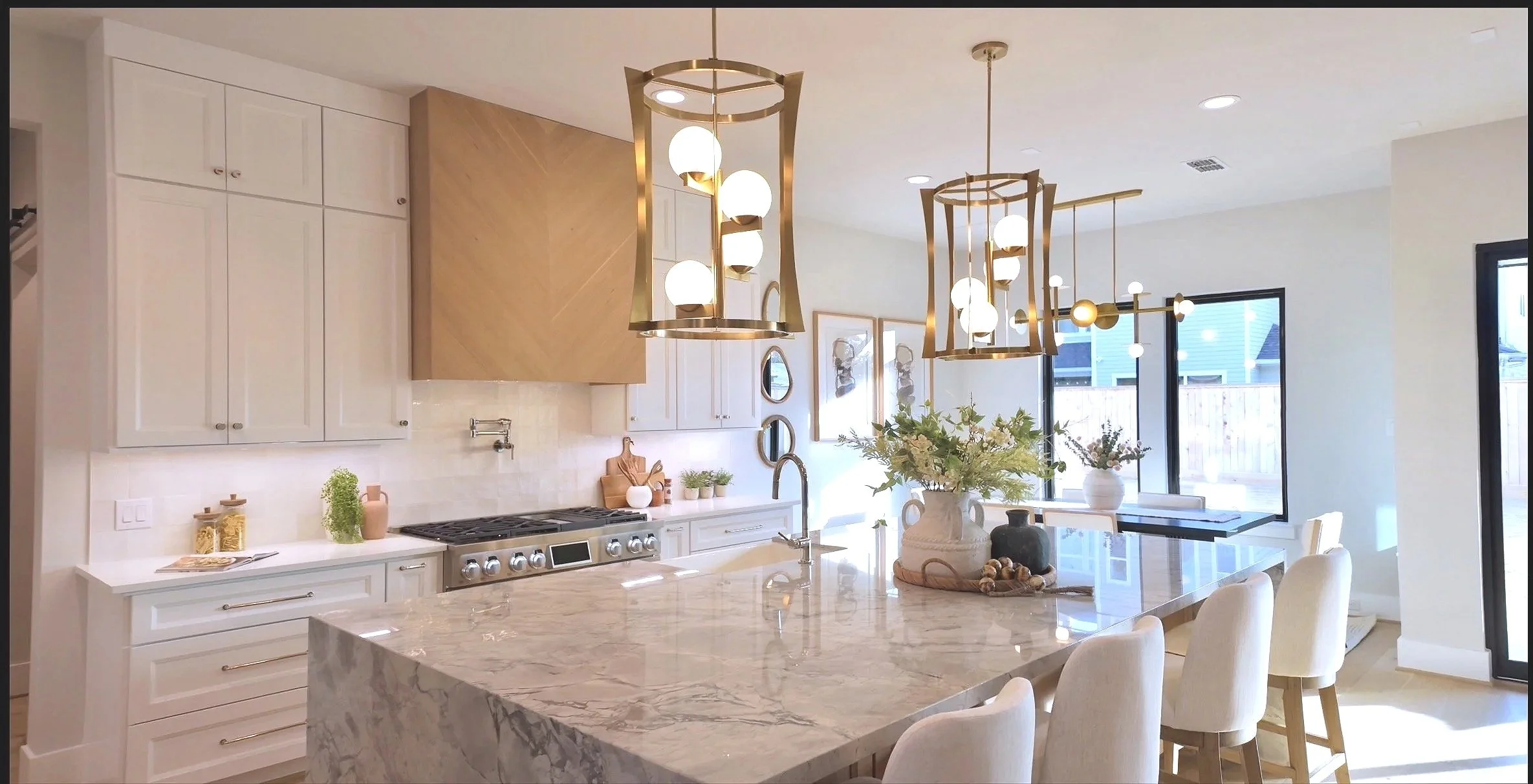 Modern kitchen with white cabinets, marble countertop, gold pendant lights, and large windows with outdoor view.