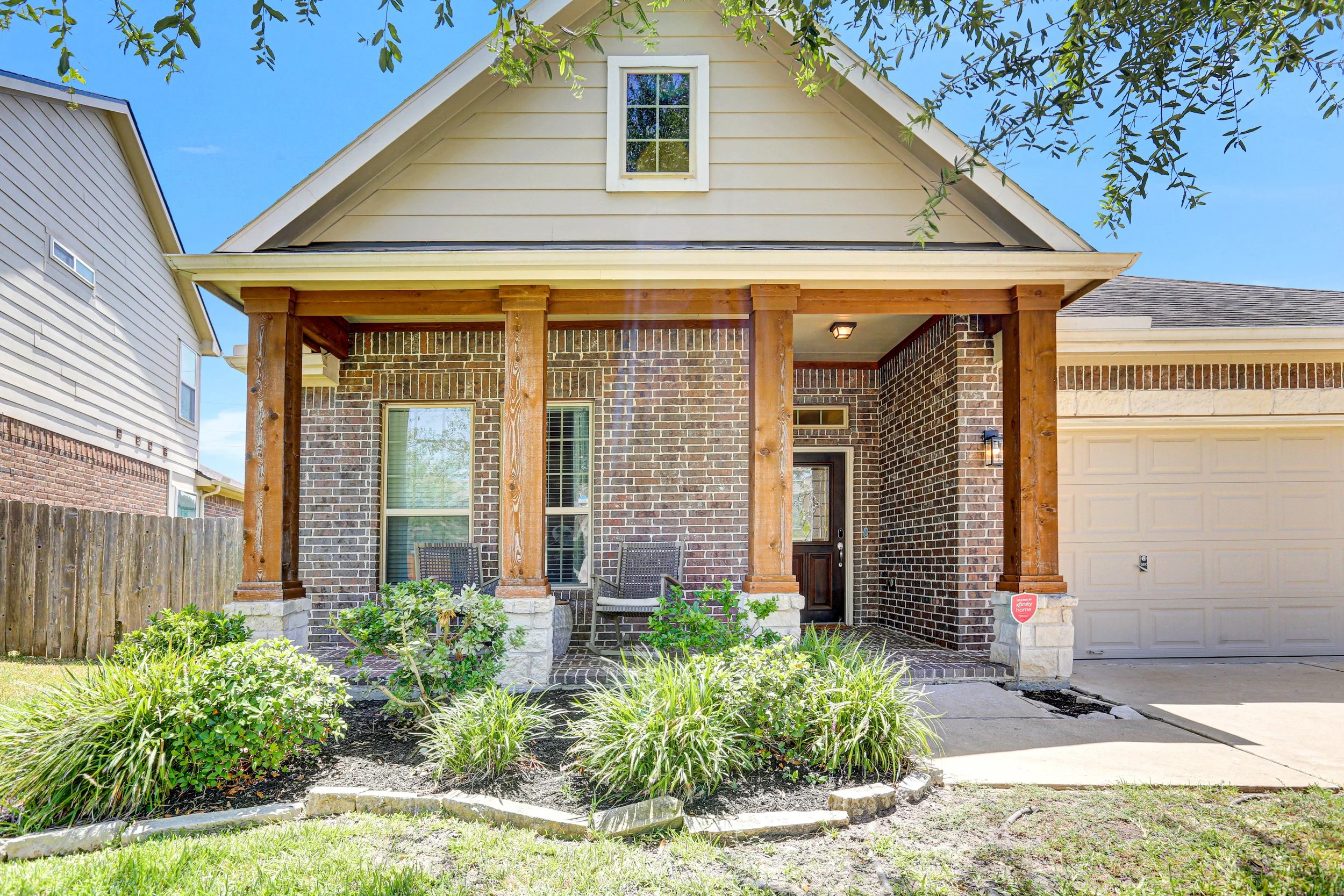 Front porch of a house with brick and siding exterior, wooden columns, two chairs, a small table, and garden plants, under a tree with blue sky in Houston