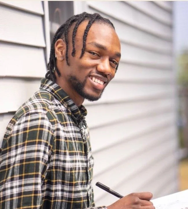 A young man with braided hair smiling while holding a pen in front of a light-colored wooden wall.
