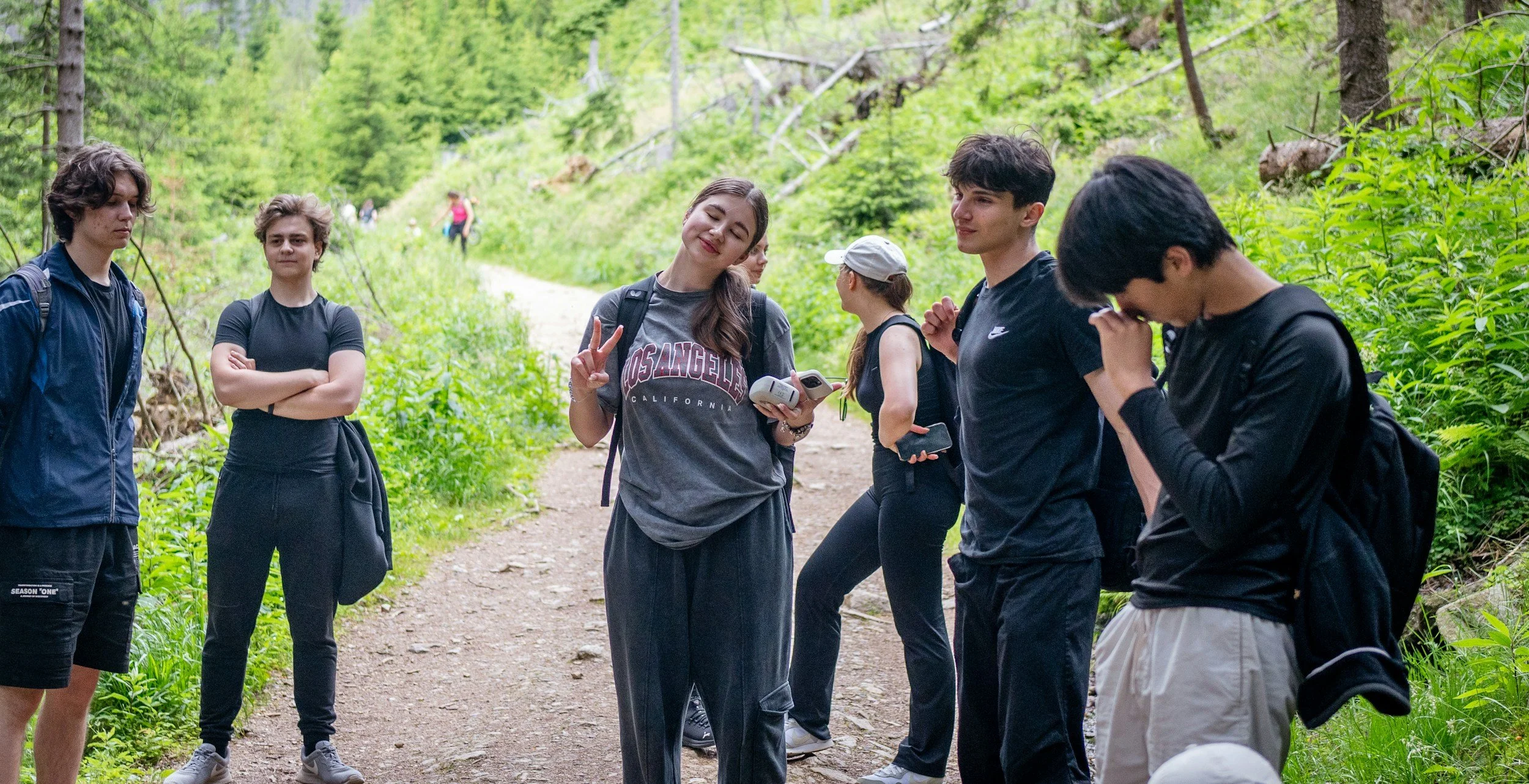 Group of young people hiking on a wooded trail, some talking, one making a peace sign, three with backpacks, surrounded by lush green foliage.