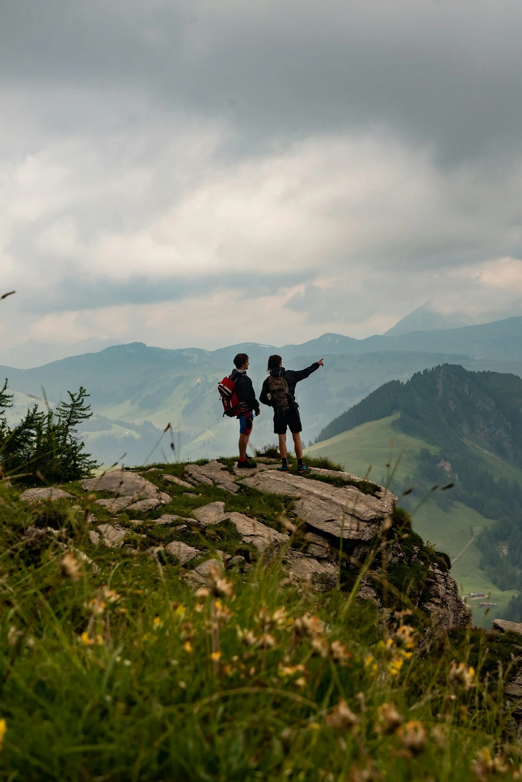 Two hikers standing on a rocky ledge overlooking mountainous landscape with cloudy sky, some snow on mountain peaks, and green valleys below.