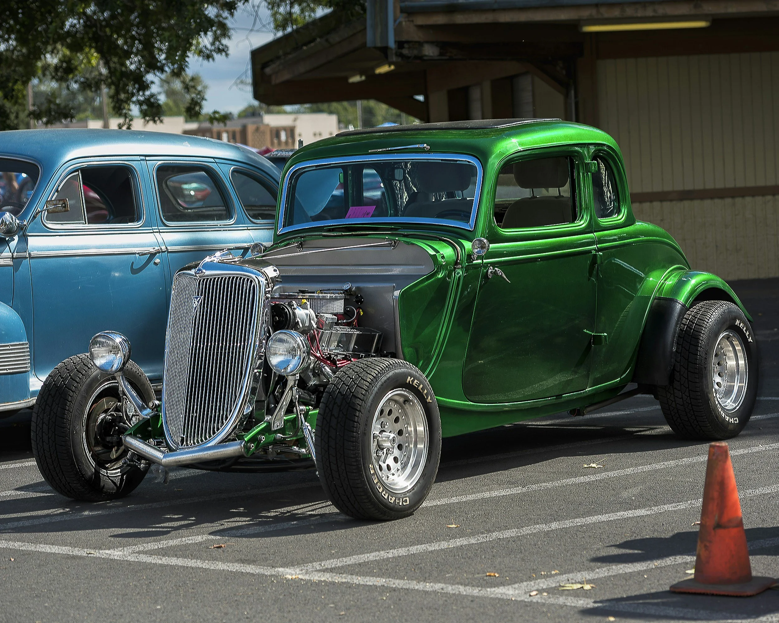 A green vintage car with chrome accents parked next to an older blue car in an outdoor lot during daytime. A traffic cone is visible in the foreground, and trees and a building are in the background.