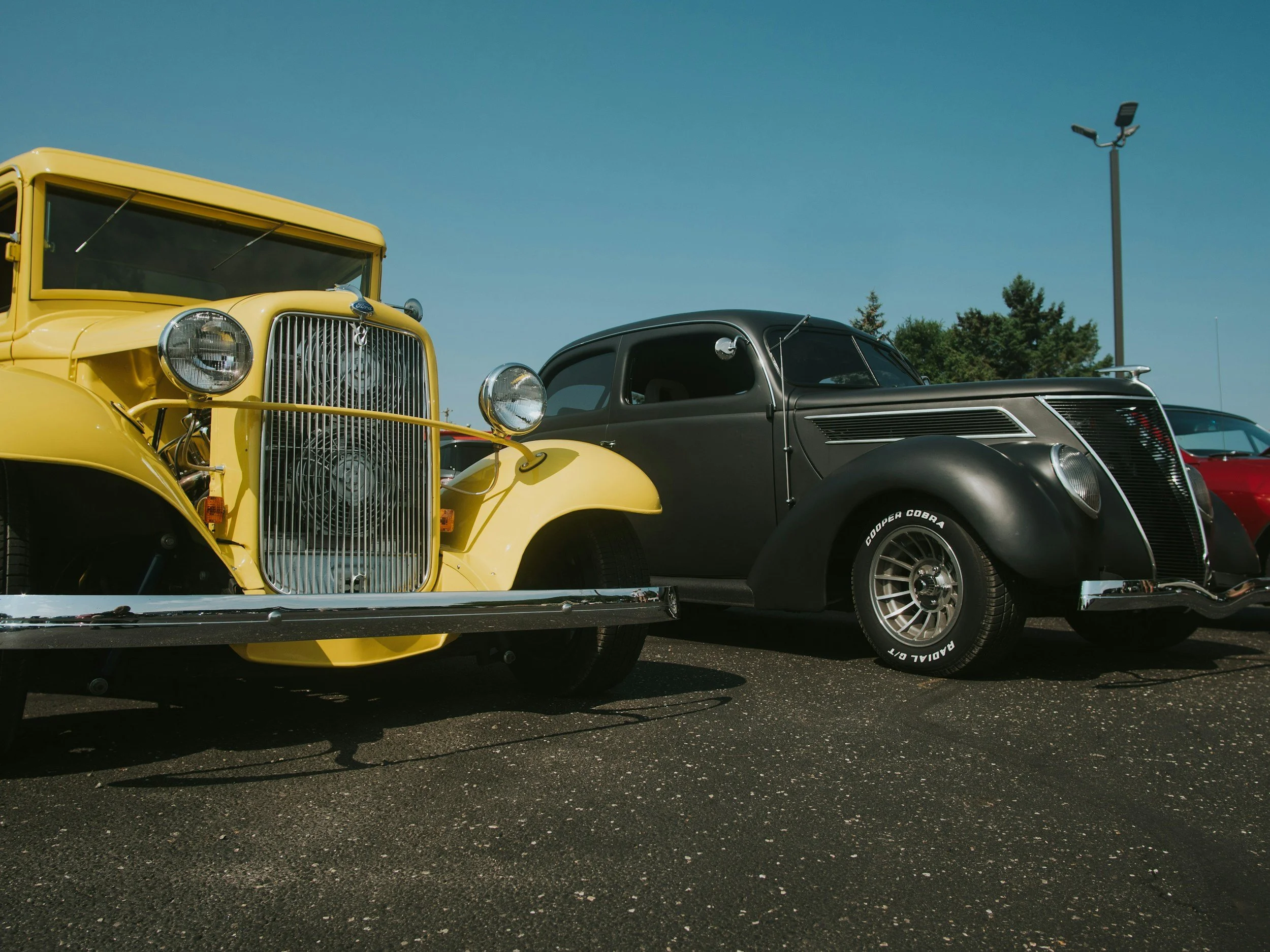 A vintage yellow car and a black classic car parked side by side in an outdoor lot, with a clear blue sky and trees in the background.