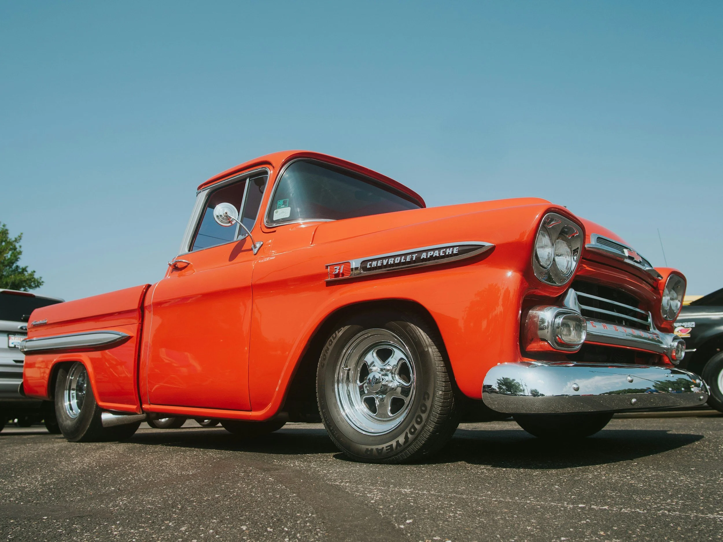 Front and side view of a vintage orange Chevrolet Apache truck parked outdoors on a sunny day with other vehicles in the background.