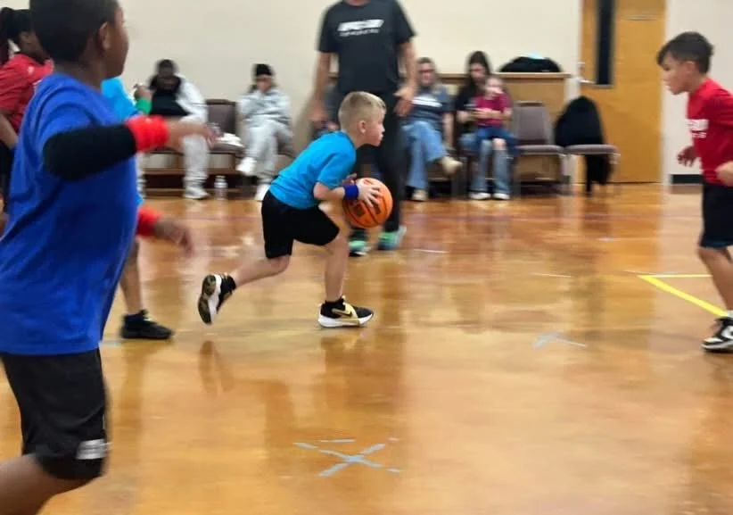Young children playing basketball in a gymnasium, with some spectators seated in the background.
