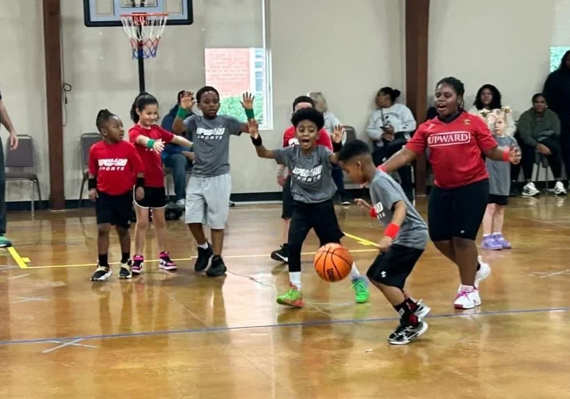 Children playing basketball indoors, with children and adults watching in the background.
