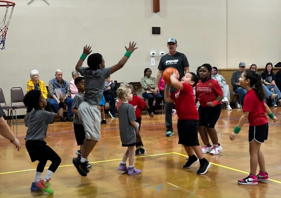 Children playing basketball in an indoor gym while spectators watch from the benches.