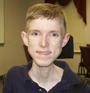 A young boy with short blond hair smiling, wearing a dark collared shirt, sitting indoors with chairs in the background.
