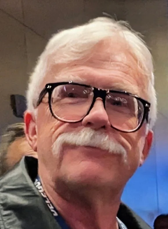 A close-up of an older man with white hair, a white mustache, wearing large black glasses, smiling slightly.