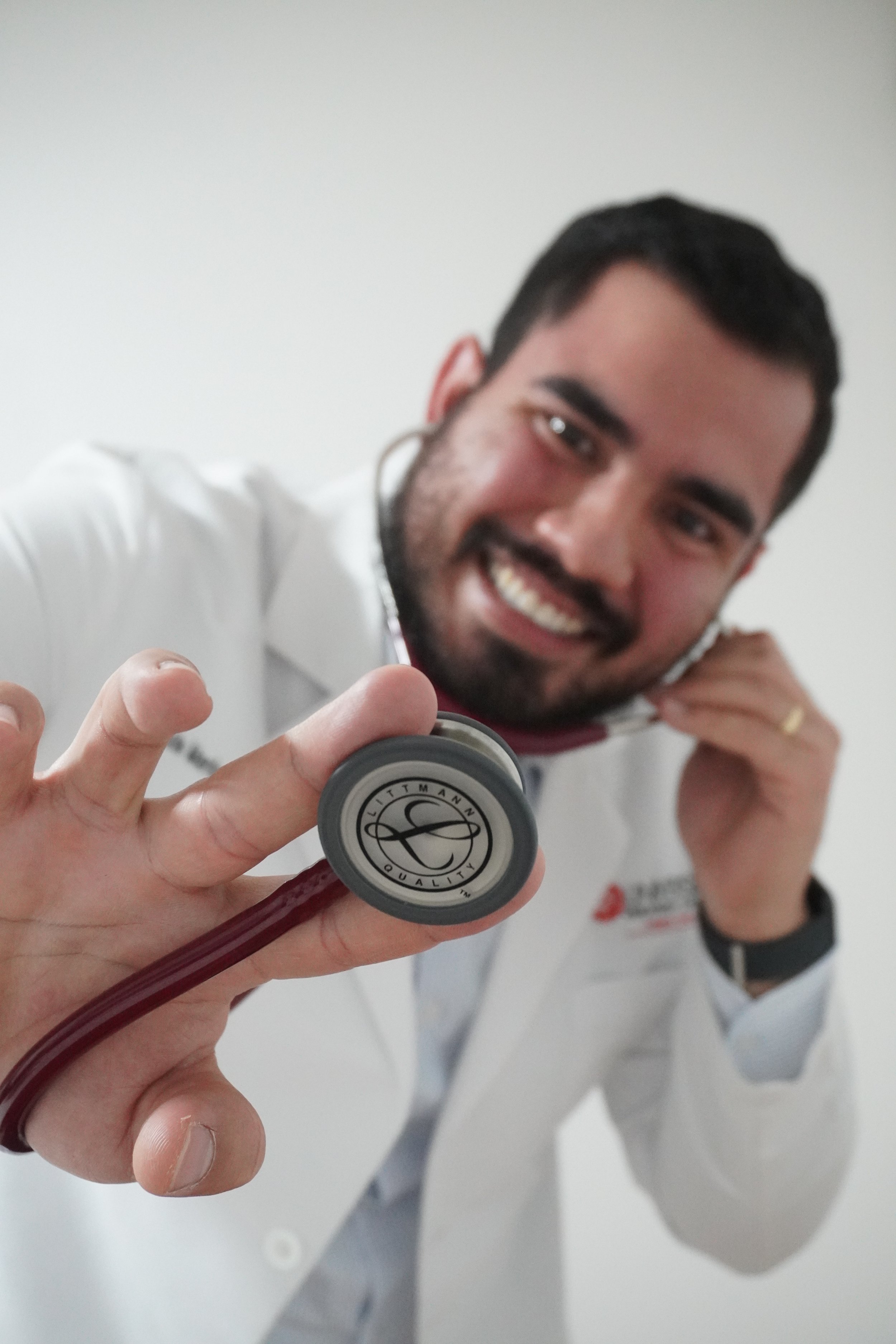 Dr. Mario Luna con bata blanca sosteniendo un estetoscopio cerca de la cámara, en un fondo blanco, consultando medicina general enfocado en estilo de vida y deporte.