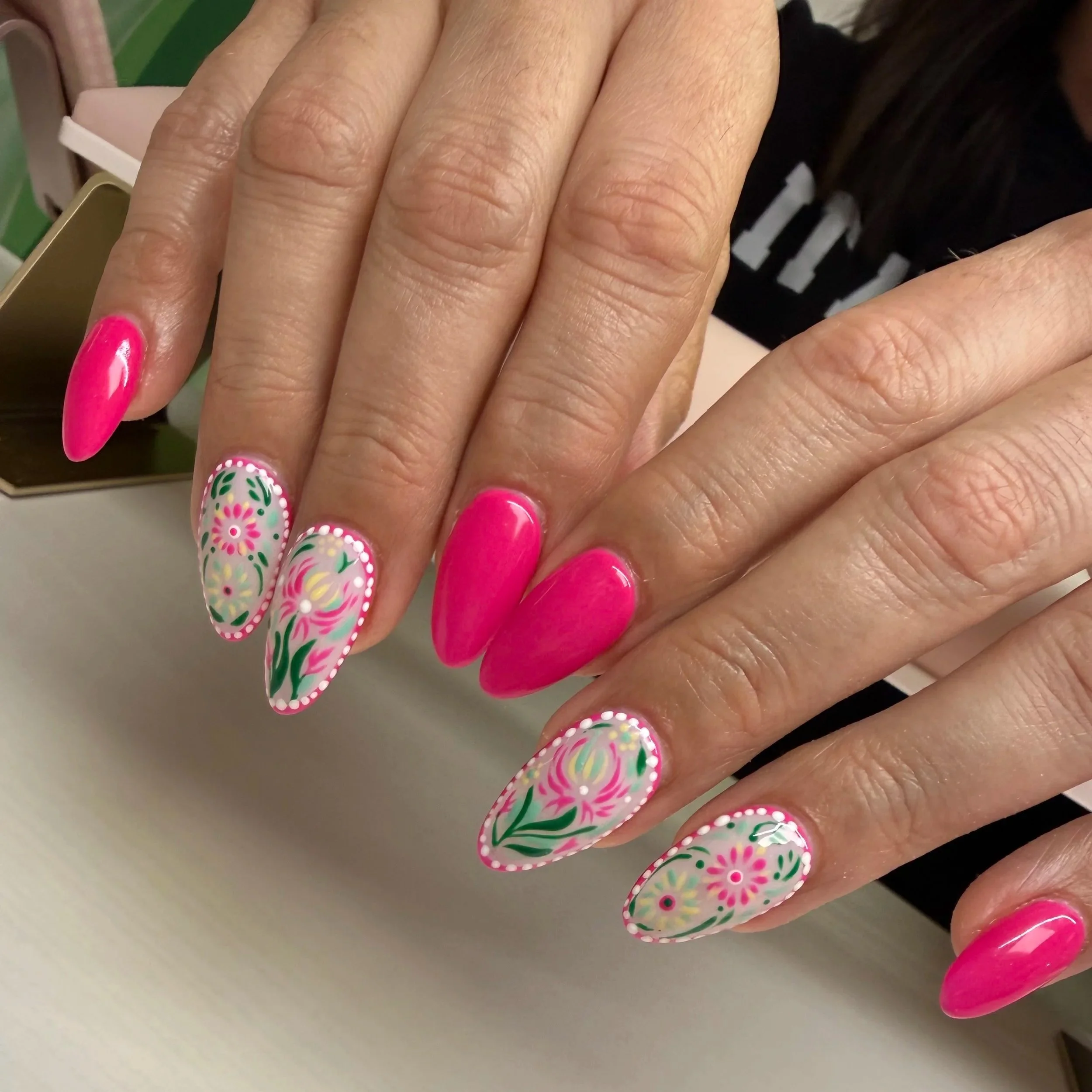 Close-up of a hand with decorated pink and floral nail polish, showing the fingers with natural and artificial nails, some painted bright pink and others with floral patterns.