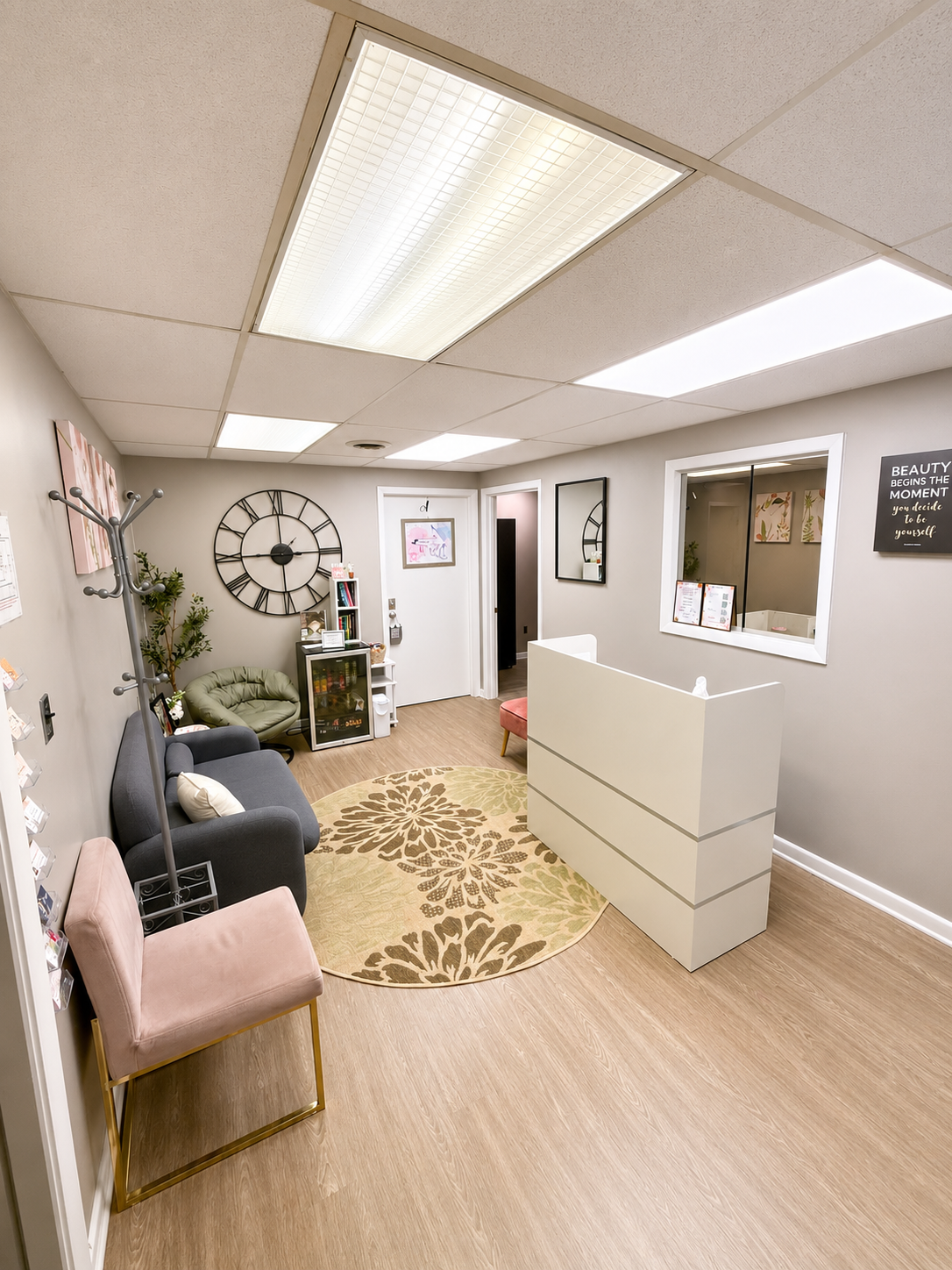 A cozy, well-lit waiting room with a round floral rug, a gray sofa with cushions, a pink chair with gold legs, a green papasan chair, a coat rack, a wall clock, framed pictures, a window with a computer screen, and a reception desk in the corner.