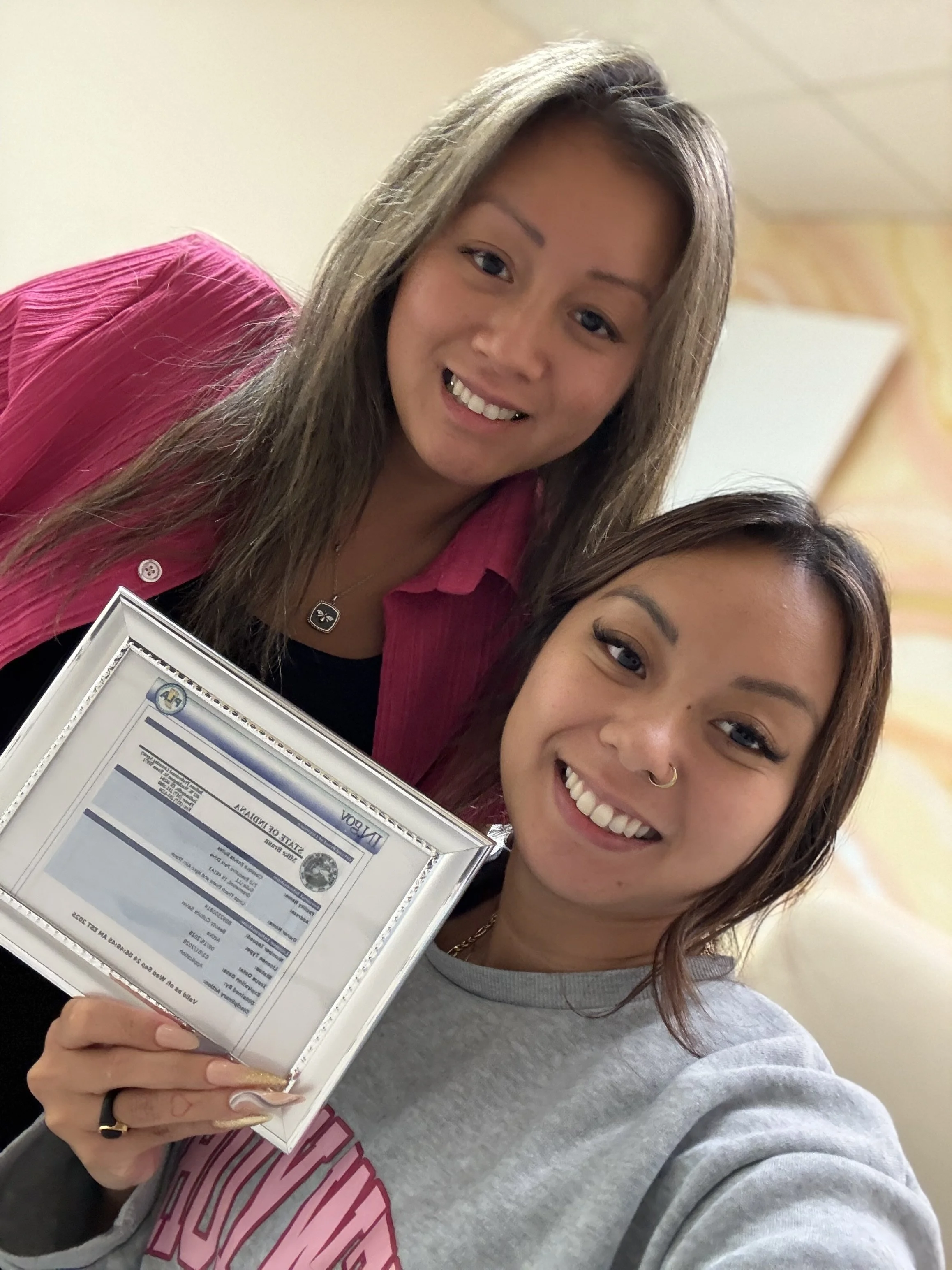 Two young women smiling, holding a framed document or artwork, taking a selfie in an indoor setting with yellow and cream walls.