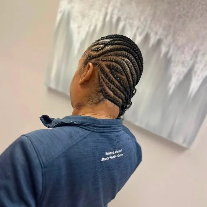 Back view of a woman with neatly braided cornrows hairstyle, wearing a blue jacket with the words 'Sandra Escarcega Mental Health Center' on the back, in a room with beige walls and abstract art hanging.