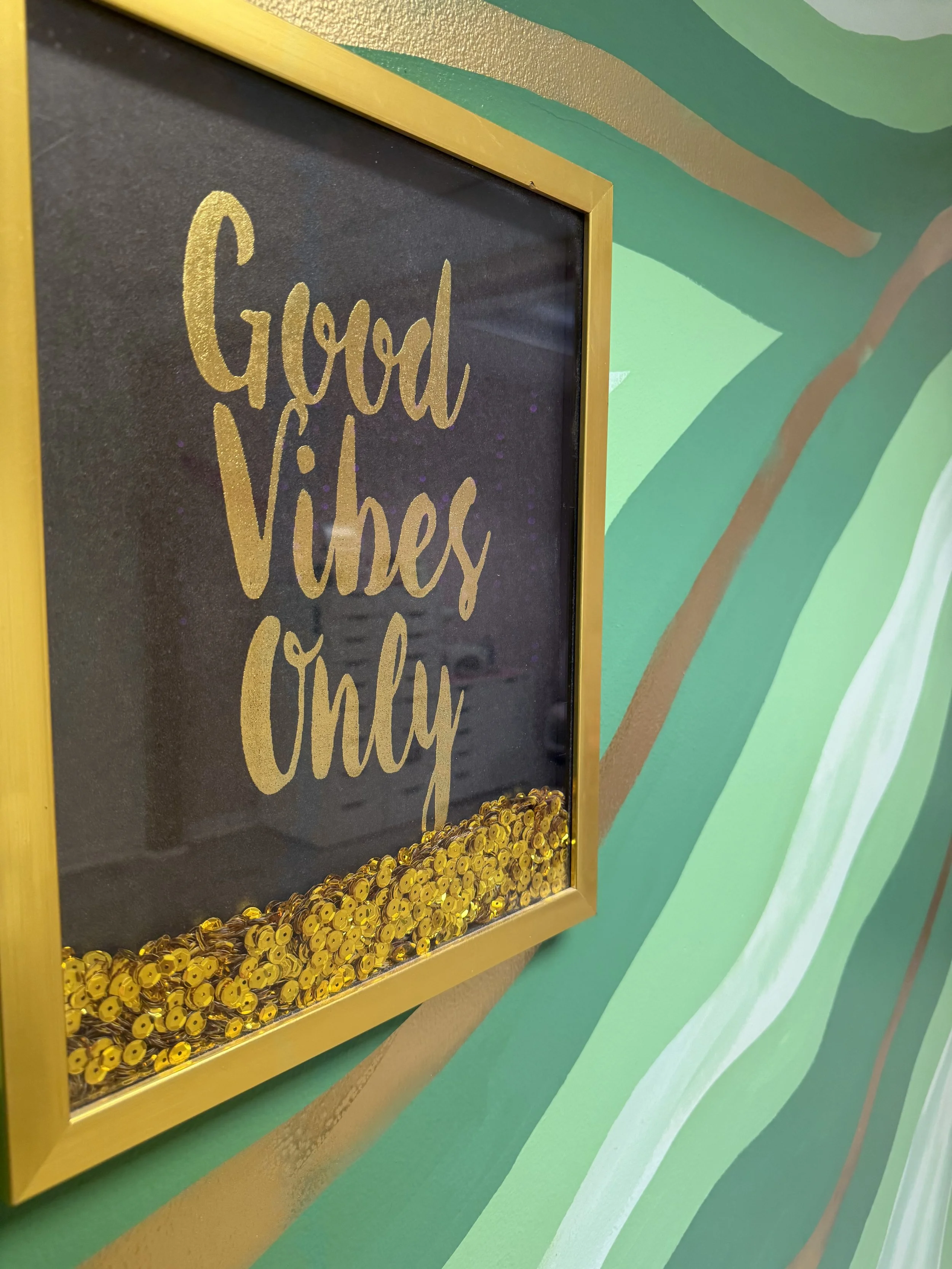 Decorative blackboard with gold-colored text that reads 'Good Vibes Only' and surrounded by gold sequins or beads, mounted on a green and gold striped wall.