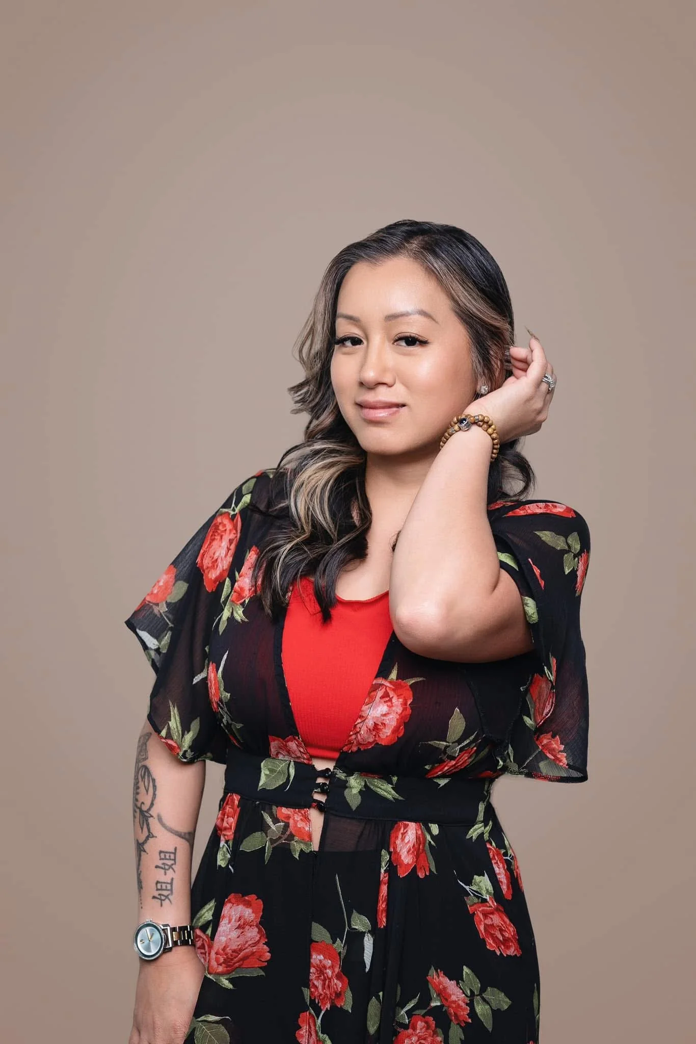 A woman with shoulder-length dark hair with blonde highlights wearing a red top and a black floral cover-up. She has a ring, a beaded bracelet, and a watch on her left wrist. She has tattoos on her left arm and is touching her hair with her right hand. The background is plain and beige.