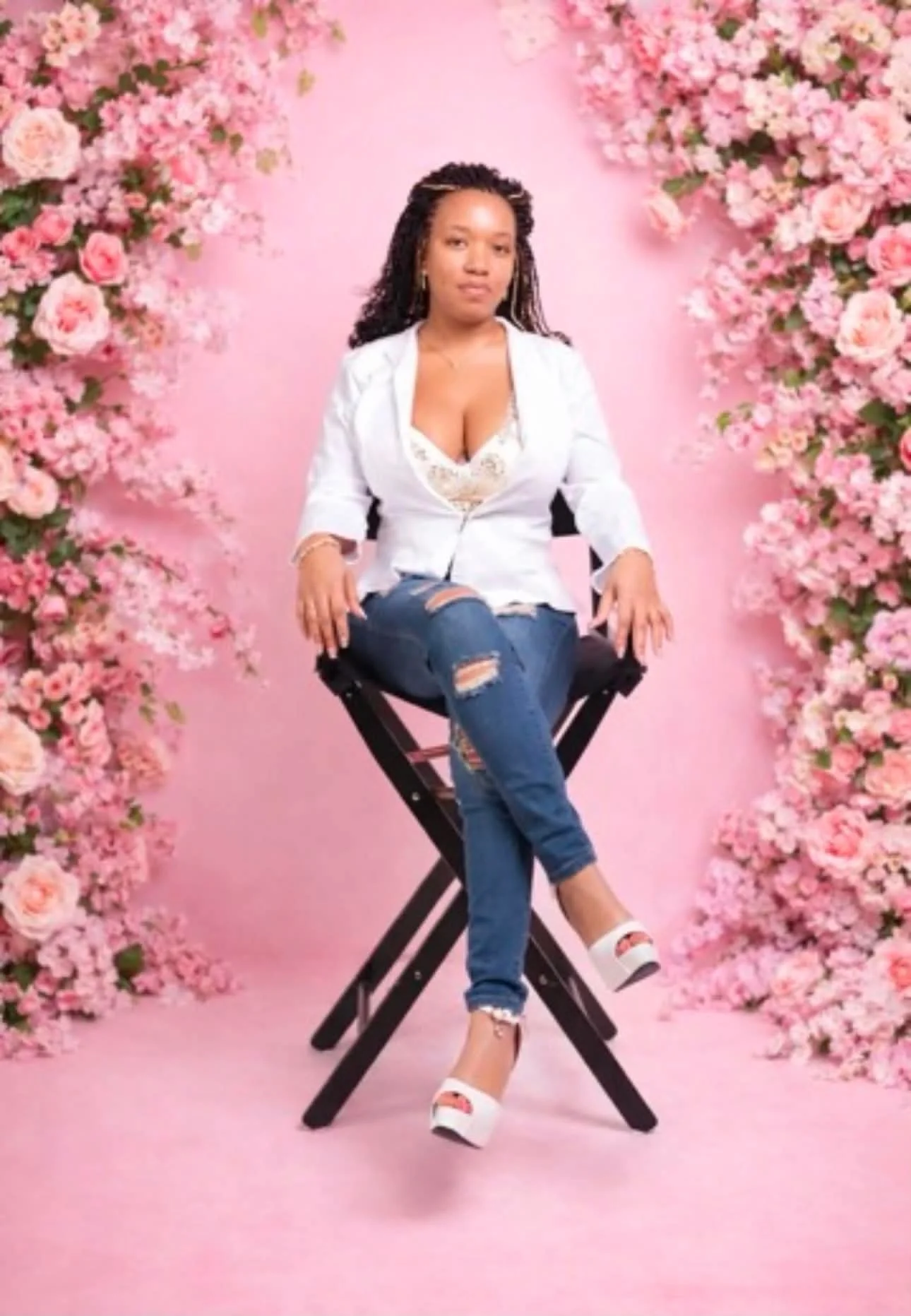 A woman sitting on a black director's chair with a pink background and large pink and white flowers on either side.