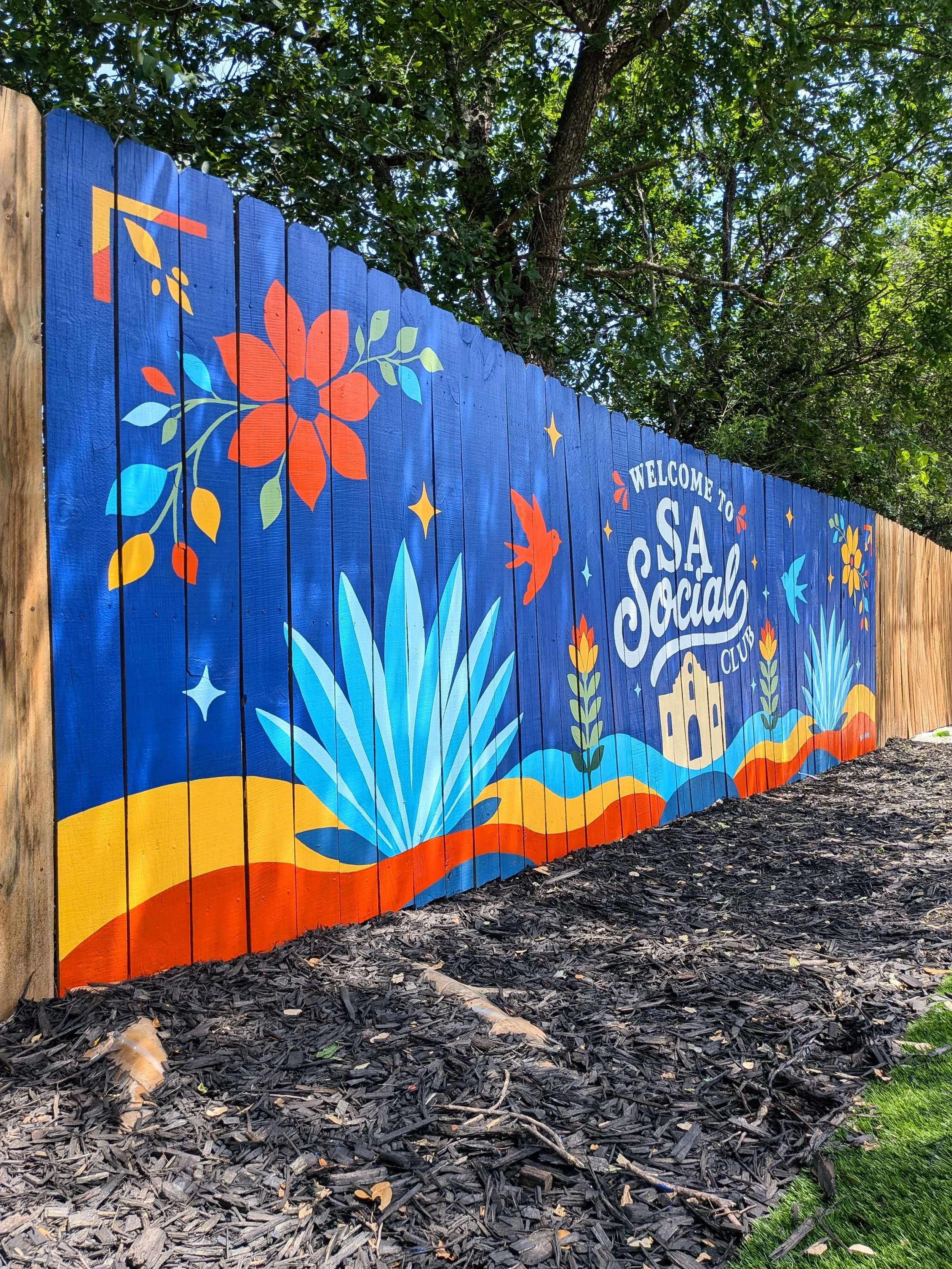 Colorful exterior fence mural for a San Antonio airbnb featuring the SA Social Club sign, orange flowers, native Texas plants, and a blue sky.