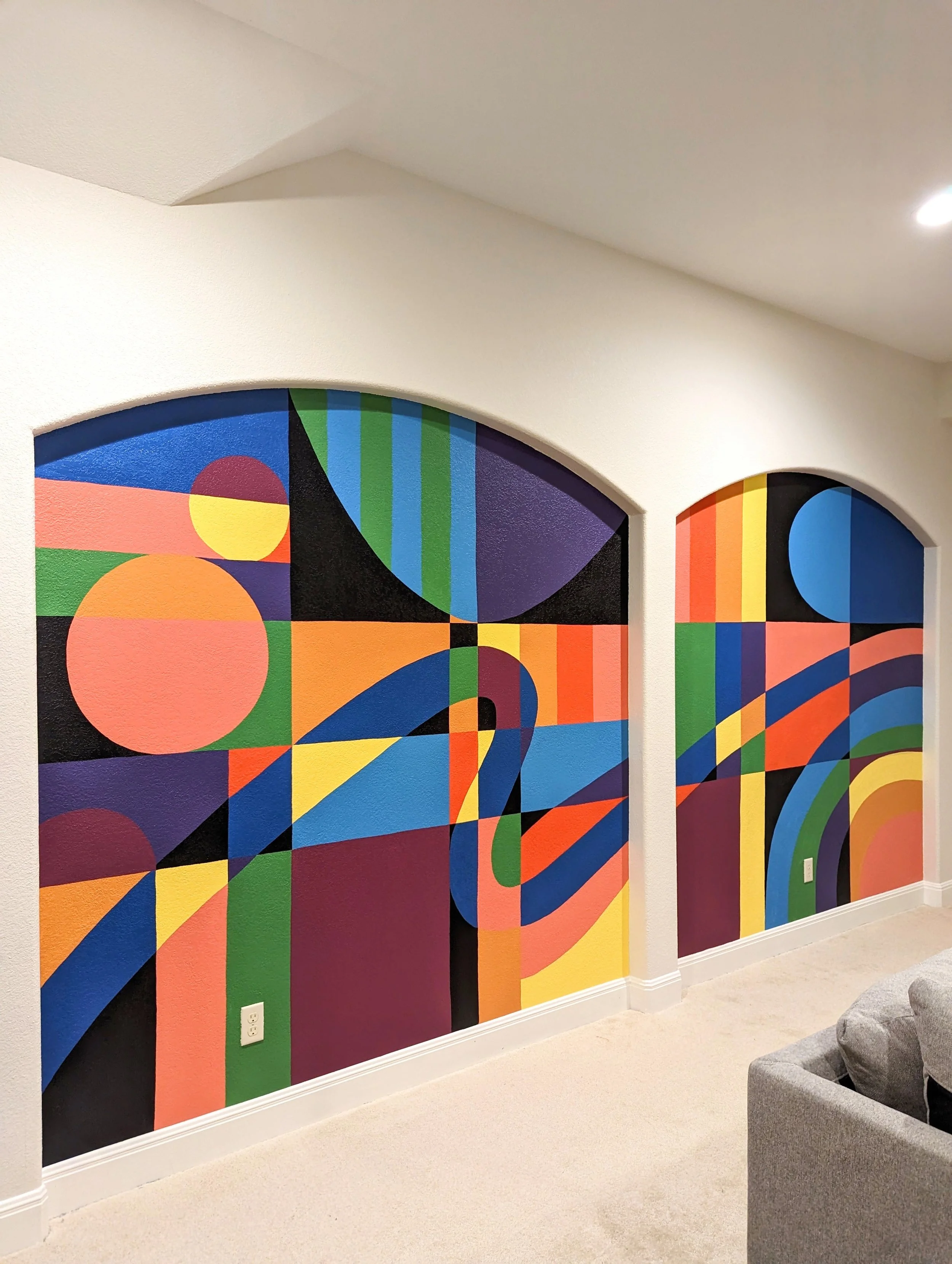 Colorful geometric murals on a wall in an interior room with beige carpeted floor and white baseboard, partially visible furniture.