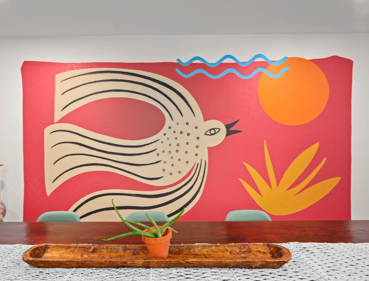 Original artwork by Tola Home featuring a folk art style bird, sun, waves, and botanical shapes in a colorful, earthy dining room.
