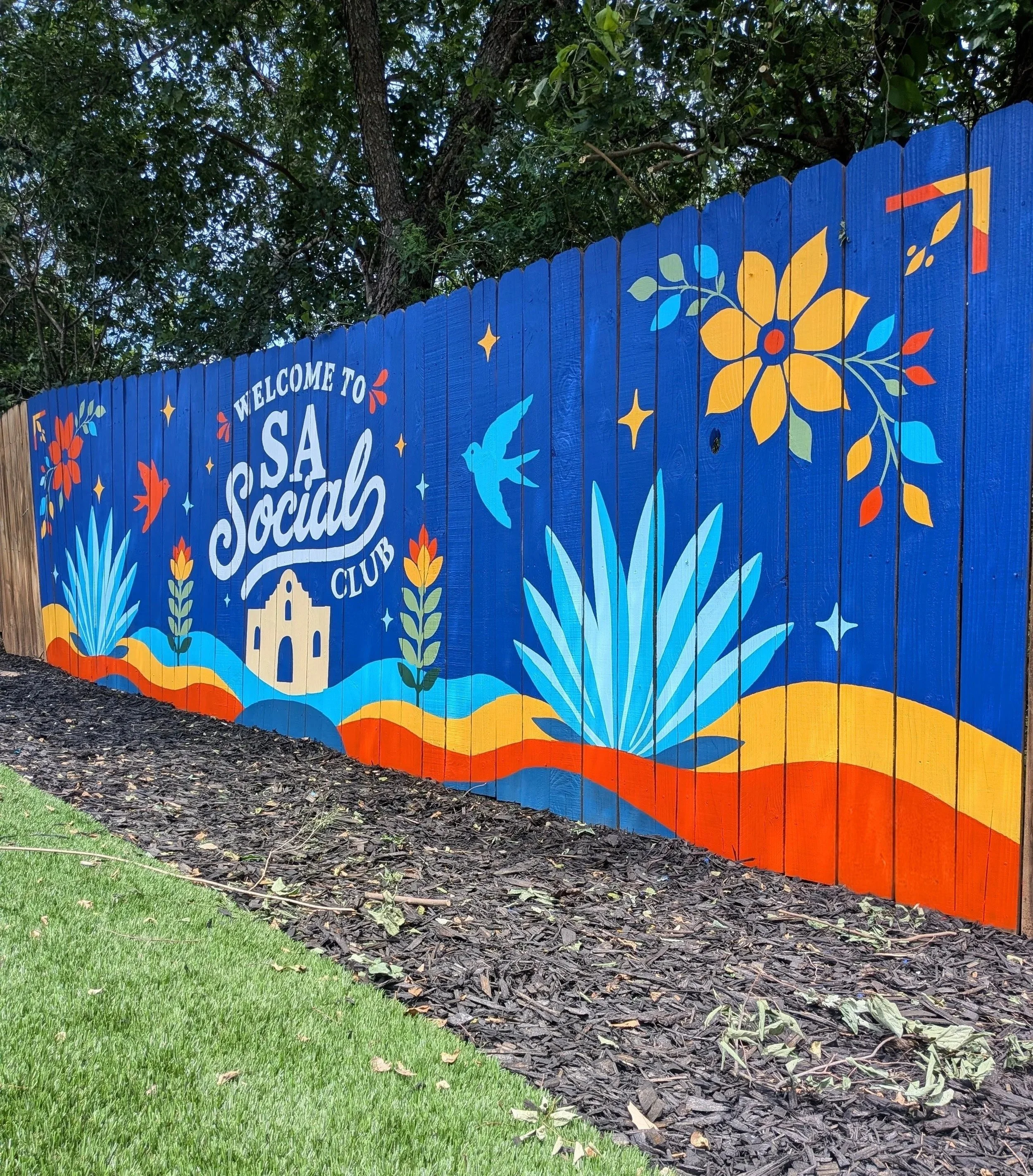 Colorful mural on a wooden fence with floral patterns, birds, stars, and the words 'Welcome to South Social Club' in white, with trees and leaves in the background.