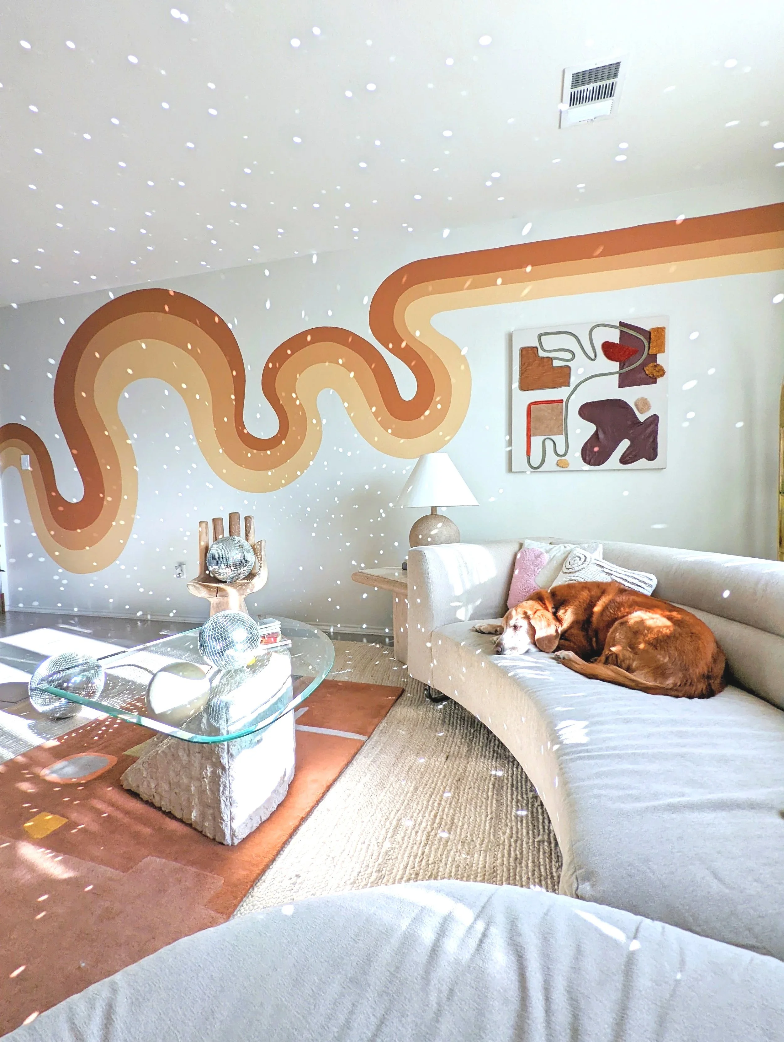 A living room with a beige sectional couch, a glass coffee table with disco balls on it, a white lamp, a colorful abstract painting on the wall, and a sleeping brown dog on the couch. The ceiling has a pattern of small lights.