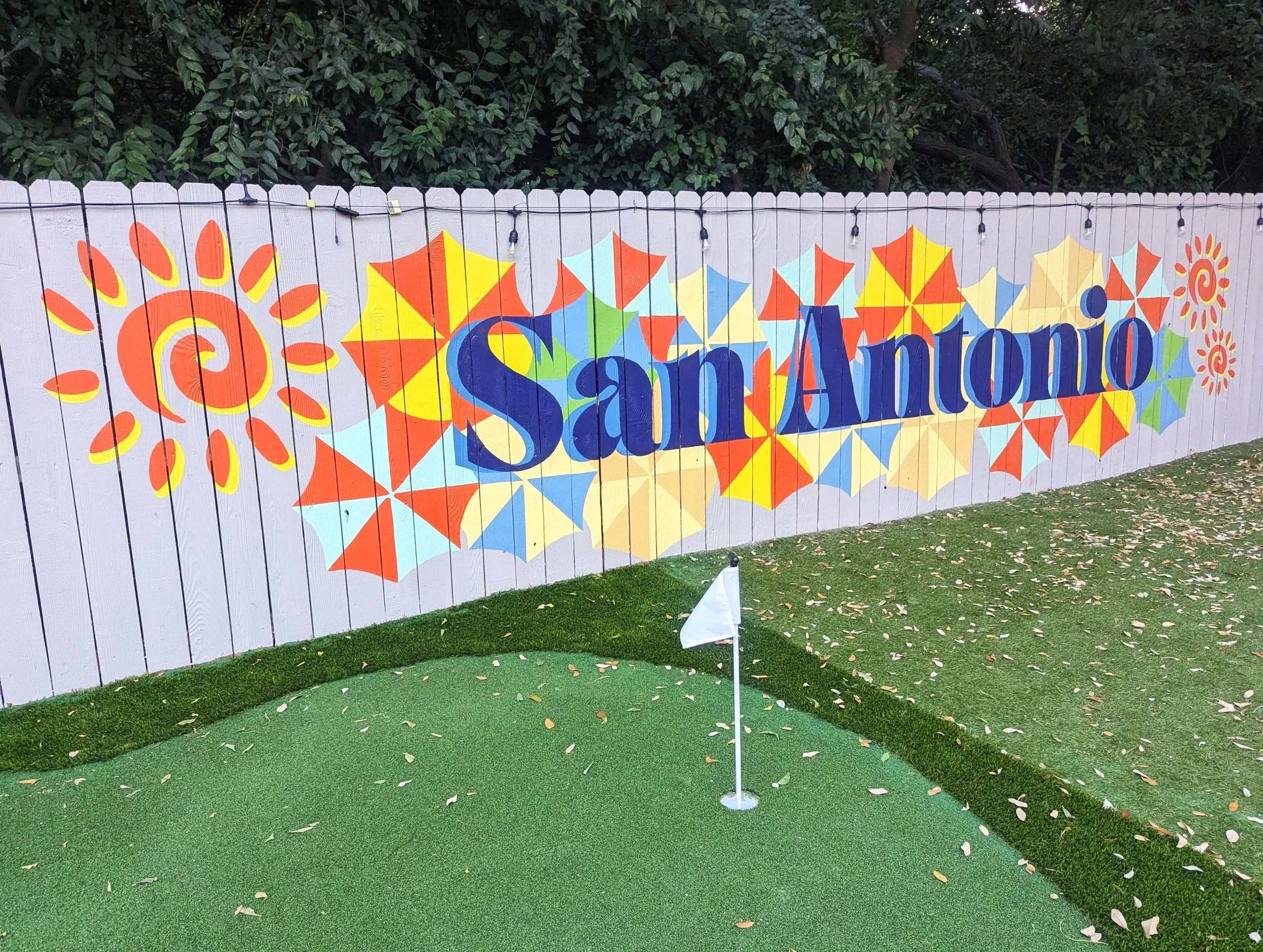 San Antonio exterior fence mural featuring blue “SAN ANTONIO” lettering over colorful fiesta umbrellas inspired by the San Antonio River Walk.