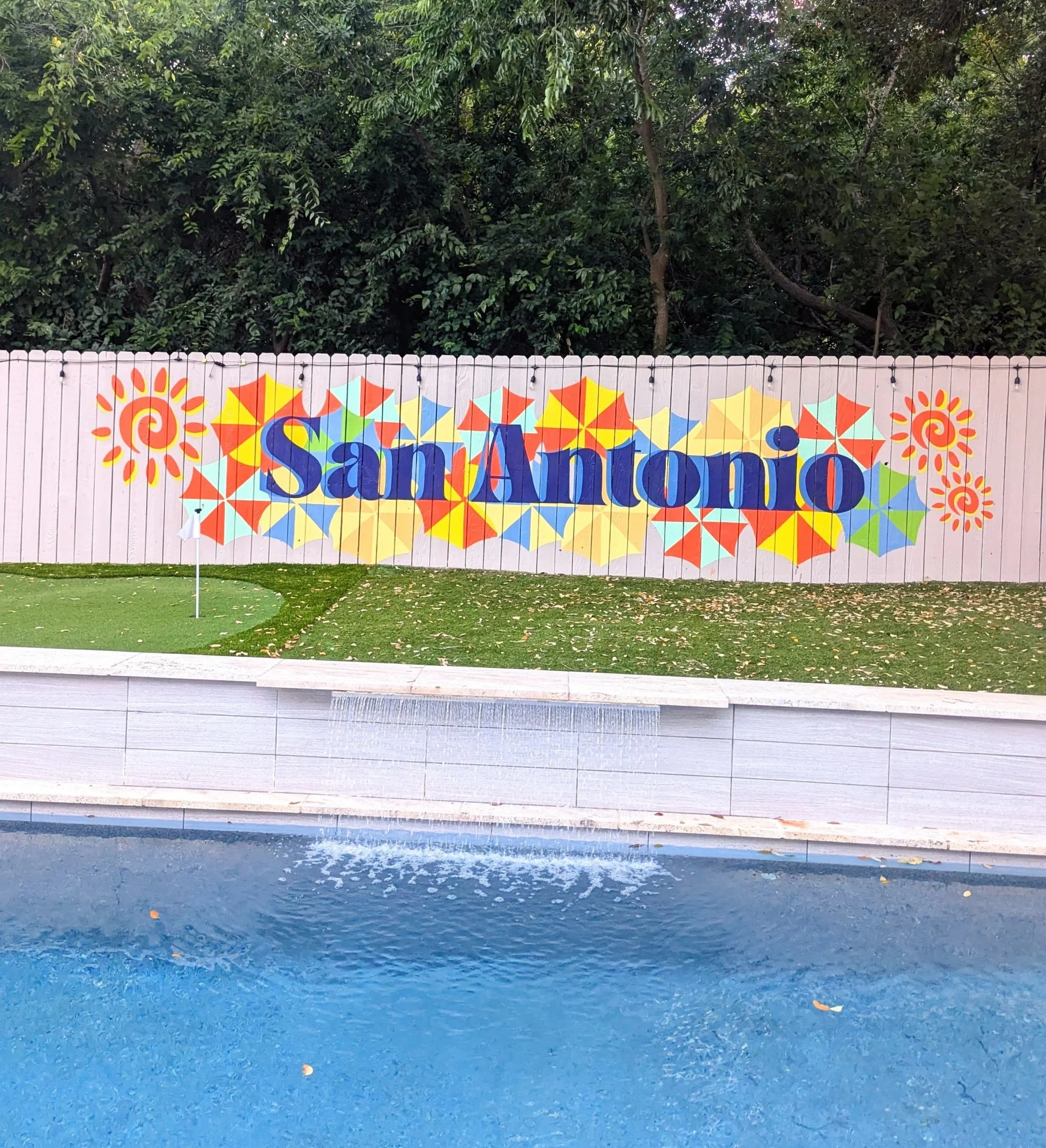 San Antonio exterior fence mural featuring blue “SAN ANTONIO” lettering over colorful fiesta umbrellas inspired by the San Antonio River Walk.