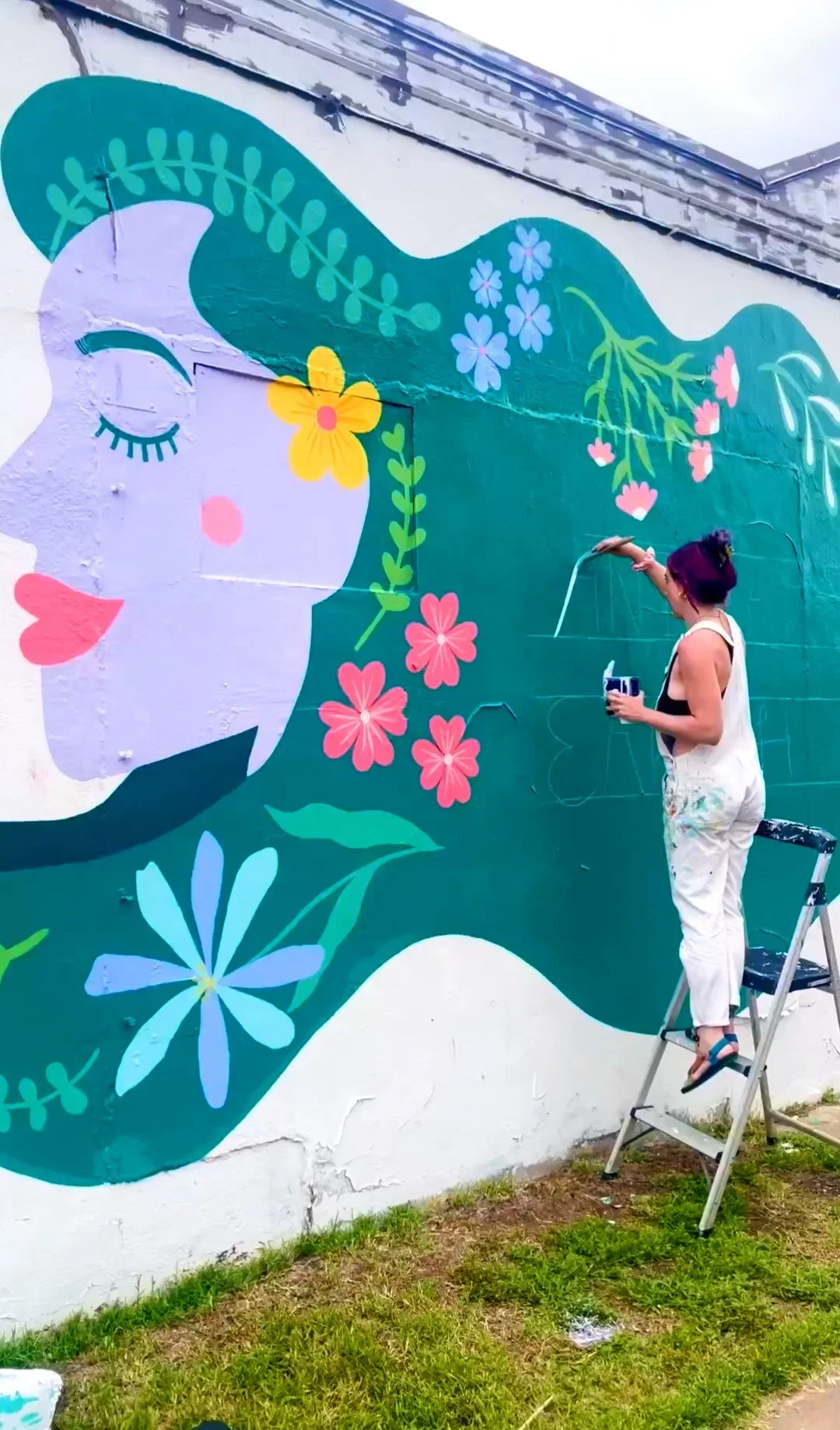 Austin Pets Alive! mural in Austin, Texas being painted on a large exterior wall with flowers, foliage, and a woman's face in style of mother earth. 
