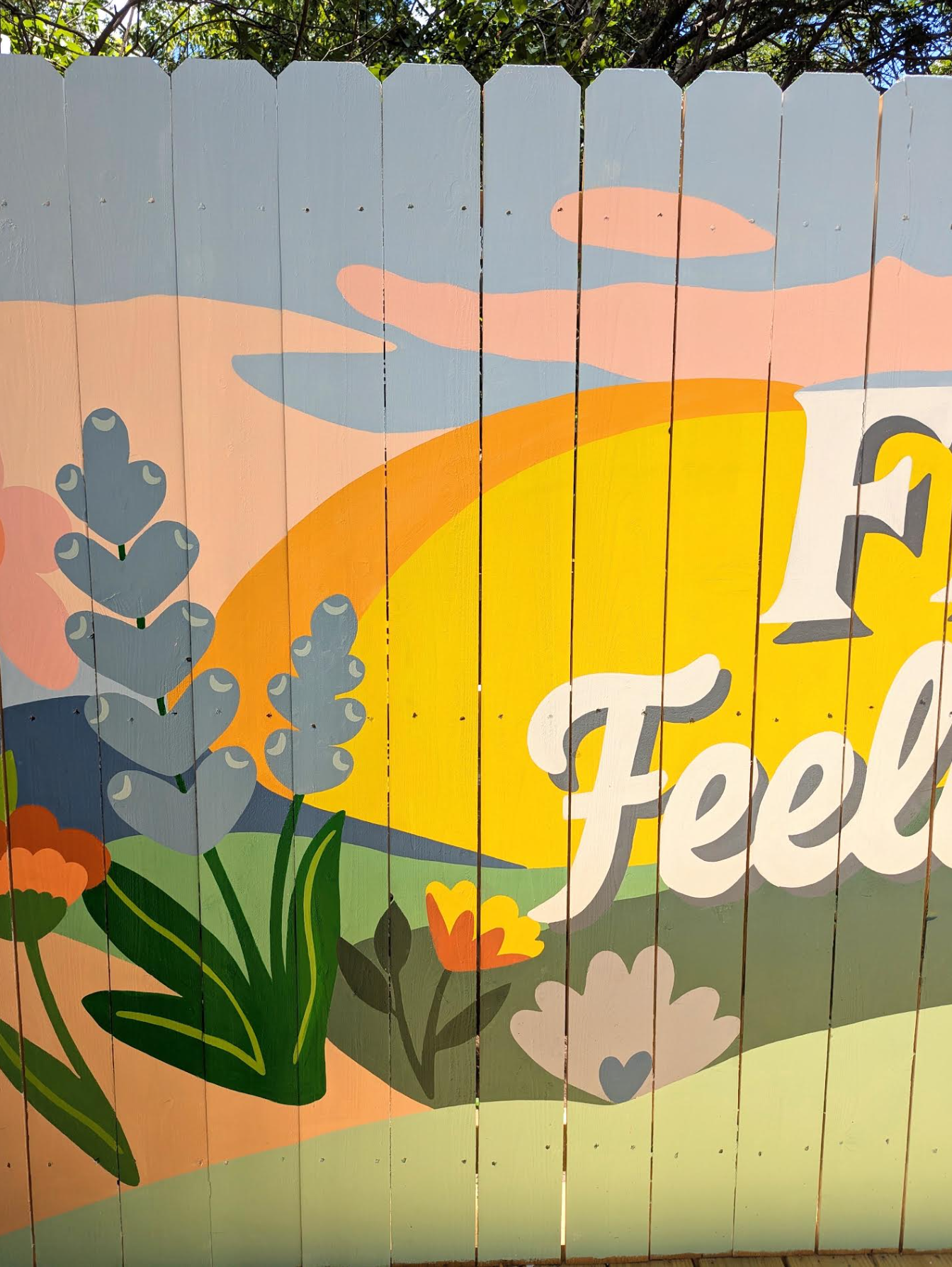 Fredericksburg, Texas short-term rental exterior fence mural featuring painted wildflowers, Texas hills, clouds, a sunset, and the phrase “FBG & Feelin' Free.”