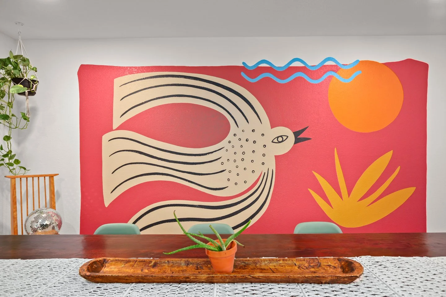 Original artwork by Tola Home featuring a folk art style bird, sun, waves, and botanical shapes in a colorful, earthy dining room.